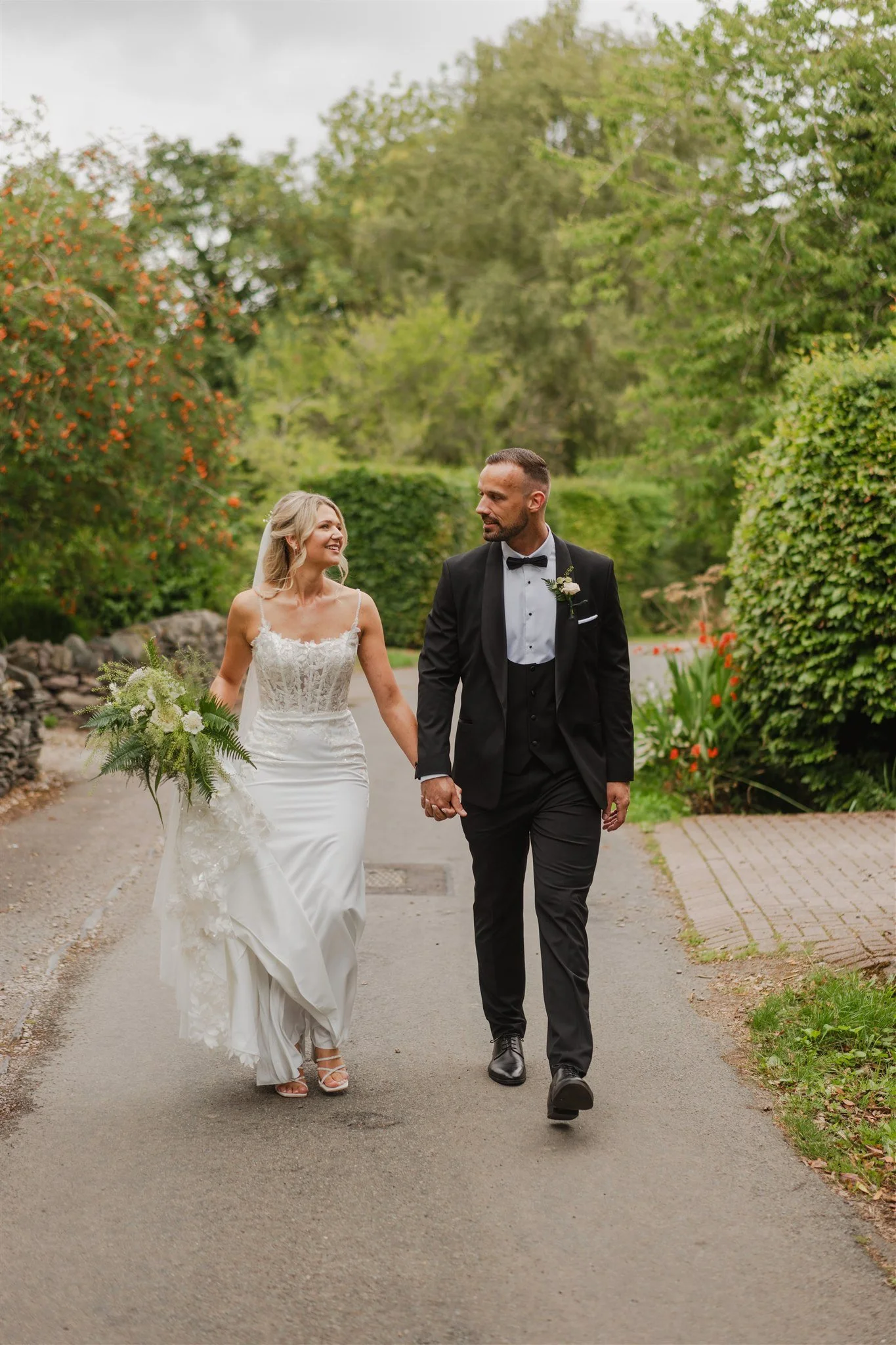 Bride and groom walking hand in hand outdoors in a park, surrounded by green trees and bushes, on their wedding day.