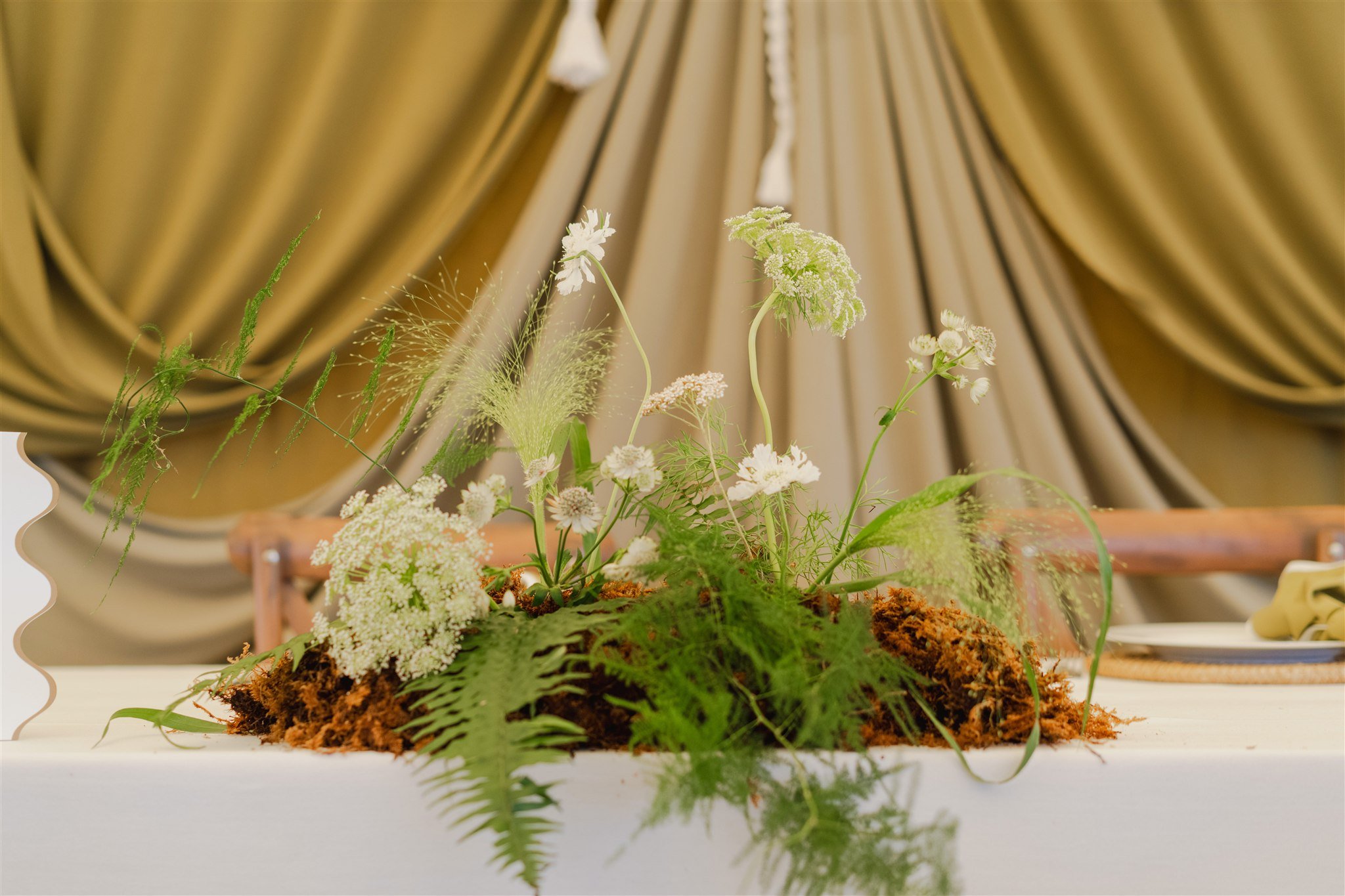 Arrangement of white flowers and greenery on a table with beige drapes in the background.