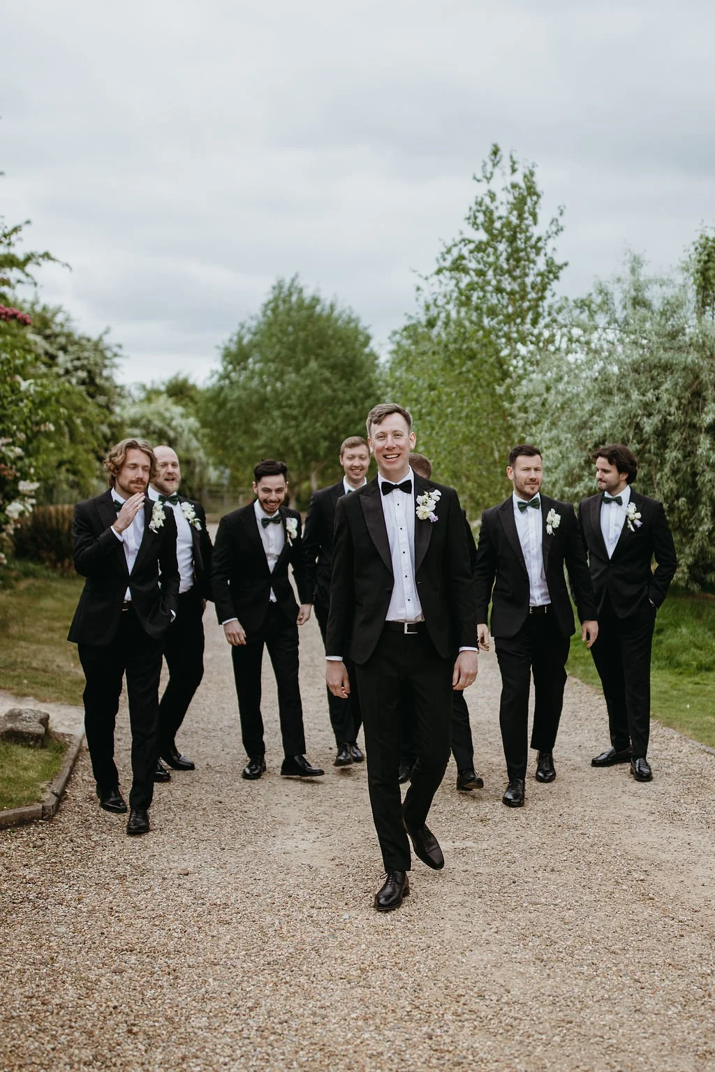 Group of men dressed in black tuxedos and bow ties walking outdoors on a gravel path, smiling, with greenery and trees in the background.