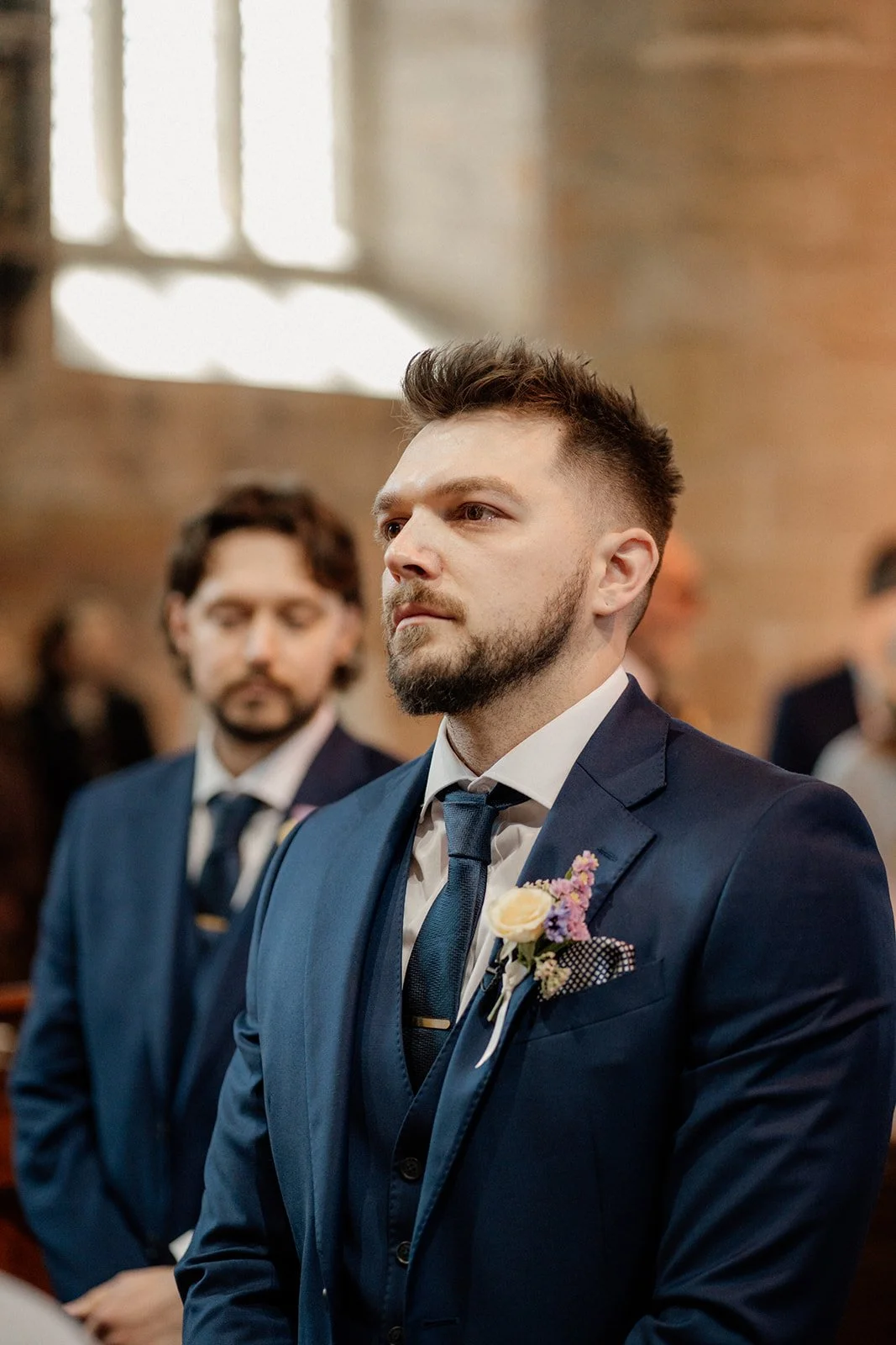 A man in a dark blue suit with a boutonniere, standing in a church during a wedding ceremony.