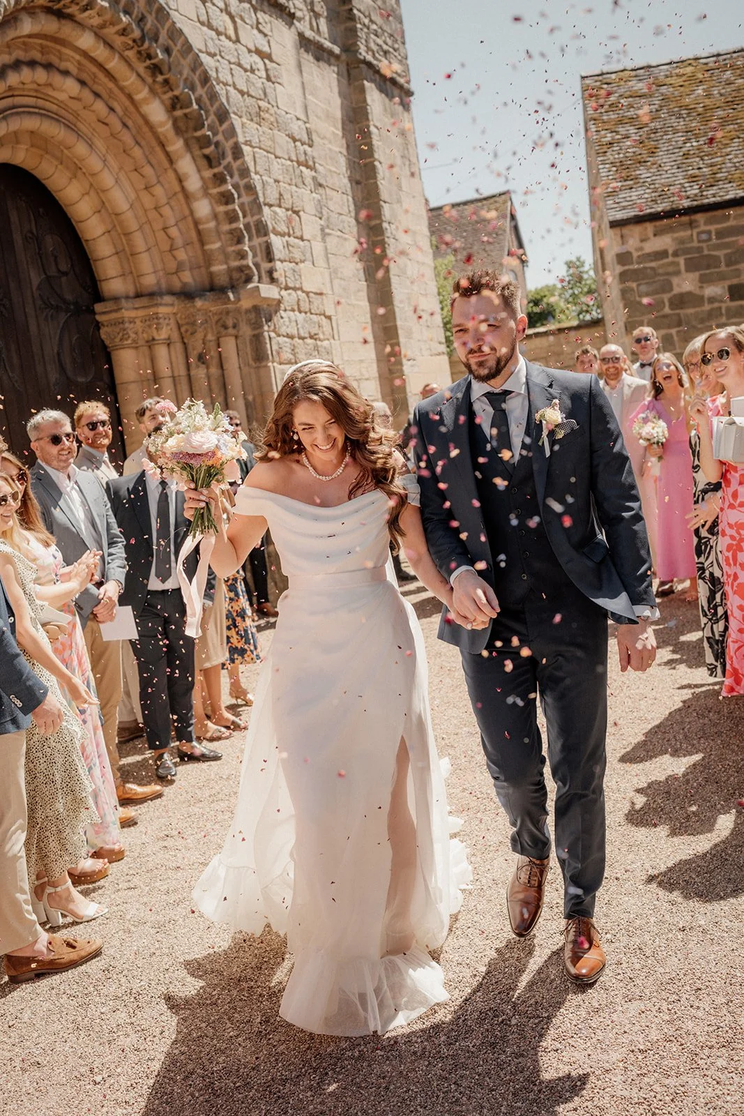 A bride and groom walking hand in hand out of a stone church, celebrating with their wedding guests who are throwing flower petals in the air.