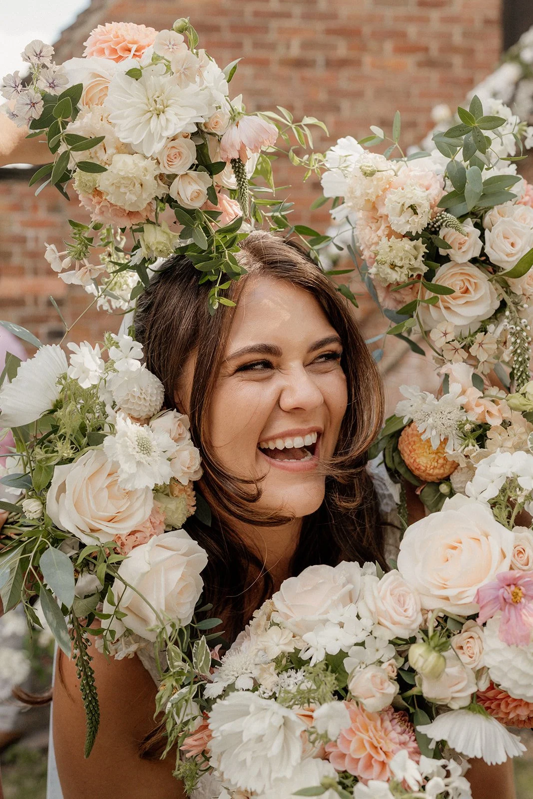 Woman smiling and laughing inside a large, colorful flower arch decorated with pastel pink, white, and peach flowers and green leaves, with a brick wall background.