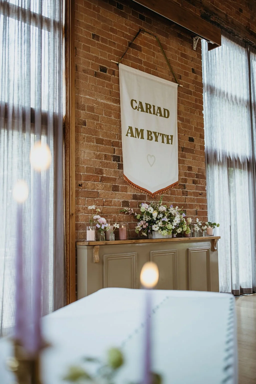 Interior of a wedding or event venue with a brick wall. A white banner with gold text hangs from a wooden beam, reading 'CARIAD AM BYTH' with a small heart. Below the banner, a beige sideboard displays a floral arrangement with pink and white flowers