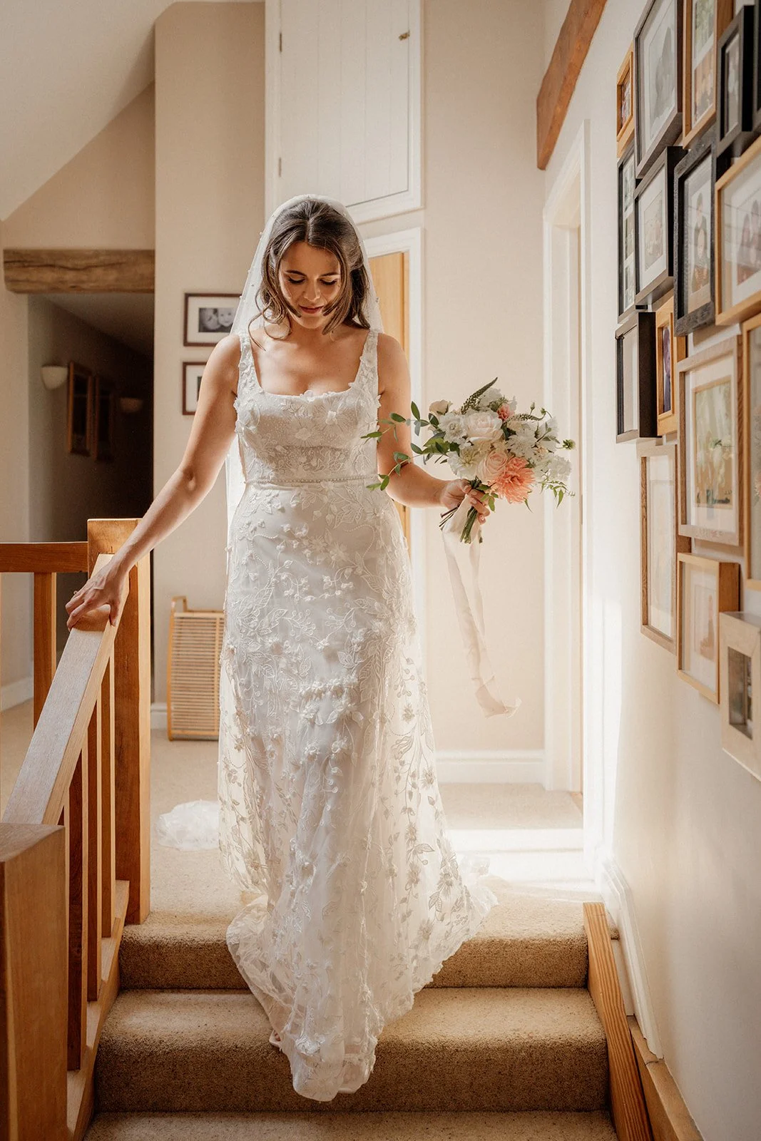 A bride in a white lace wedding dress holding a bouquet of flowers as she descends a staircase inside a house.