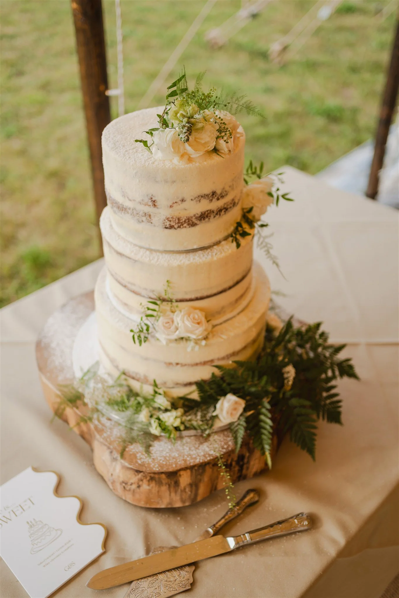 A four-tier semi-naked wedding cake decorated with white flowers and greenery, placed on a rustic wooden slab, with wedding utensils and a card nearby.