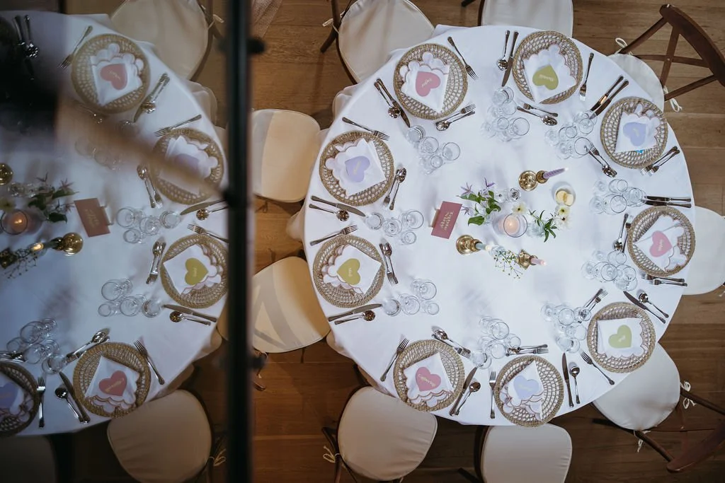 Overhead view of a round table set for a celebration with a white tablecloth, gold and white wicker chargers, colorful paper heart decorations, glasses, and silverware. The centerpiece has flowers and candles.