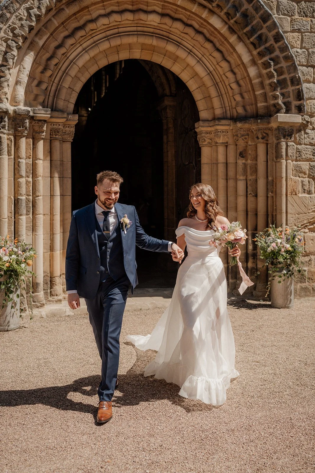 A newlywed couple walking out of a church holding hands, smiling and carrying bouquets of flowers.
