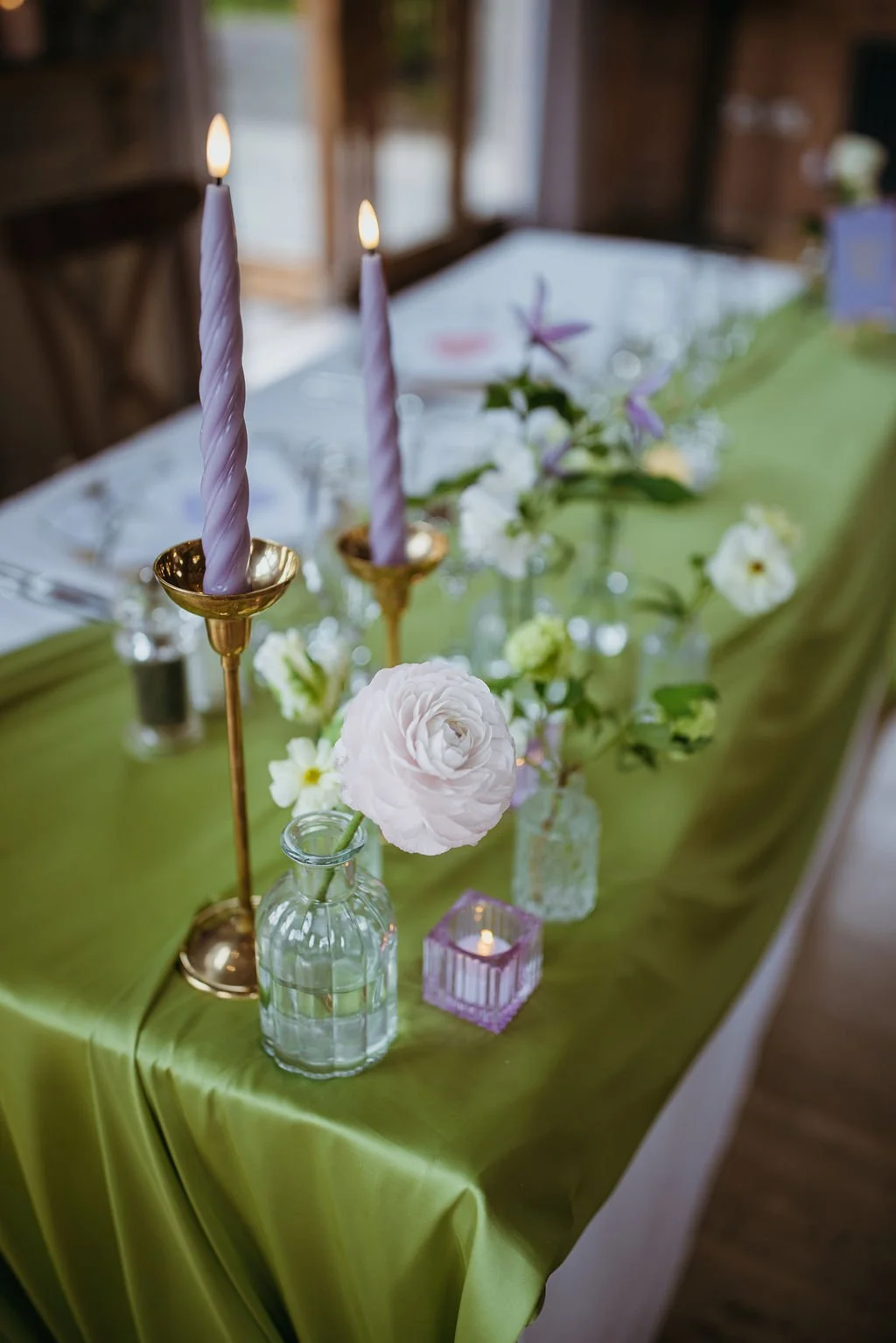A decorated table with purple candles in gold candle holders, white and purple flowers in glass jars, and a green tablecloth, set for a special occasion indoors.
