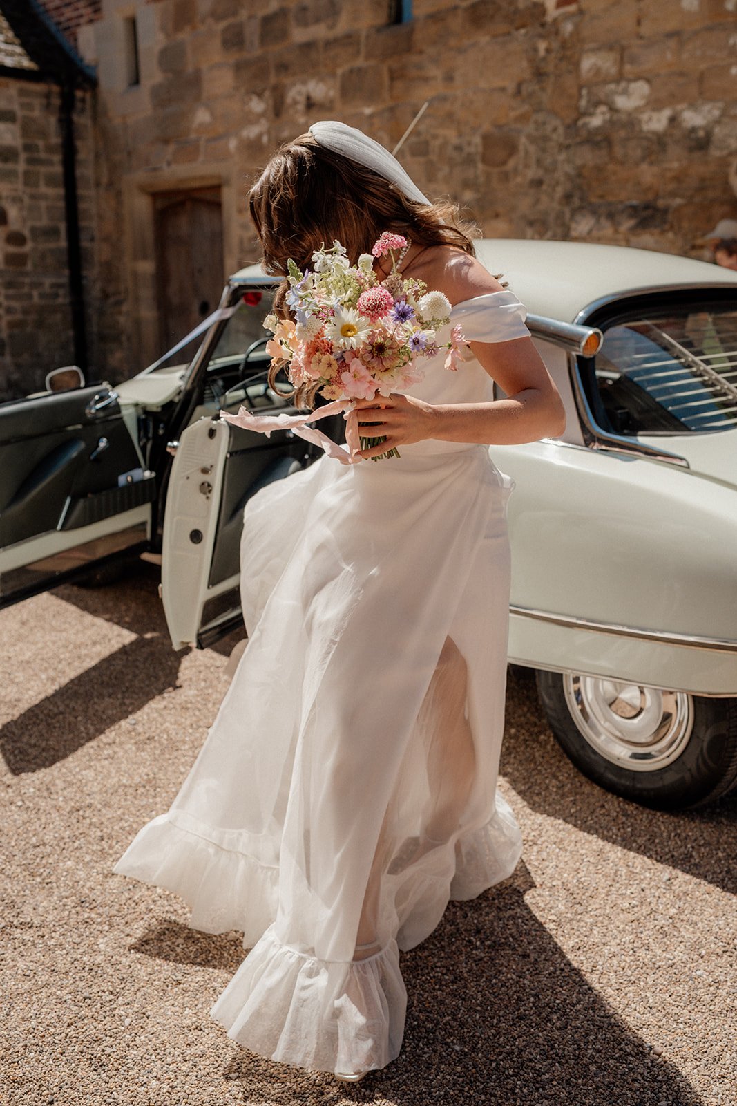 A bride in a white wedding dress holding a bouquet of colorful flowers, standing next to a vintage white car with its door open, on a gravel driveway with a brick wall in the background.