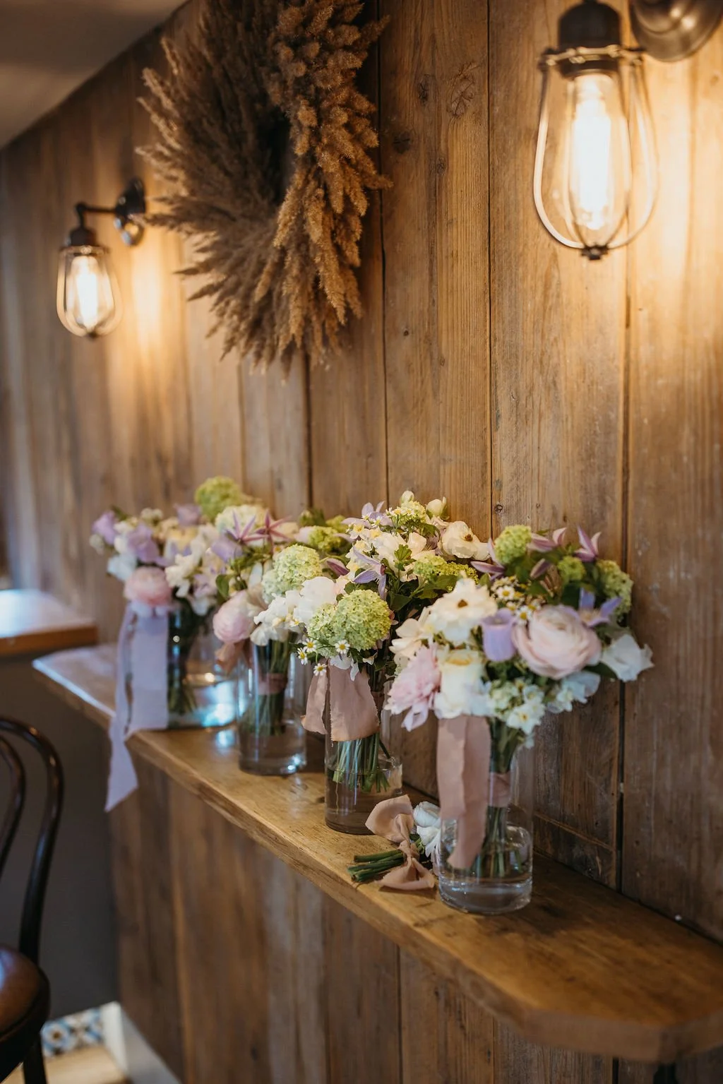 A rustic wooden shelf with several glass vases filled with pastel-colored flowers, against a wooden wall illuminated by wall-mounted light fixtures.
