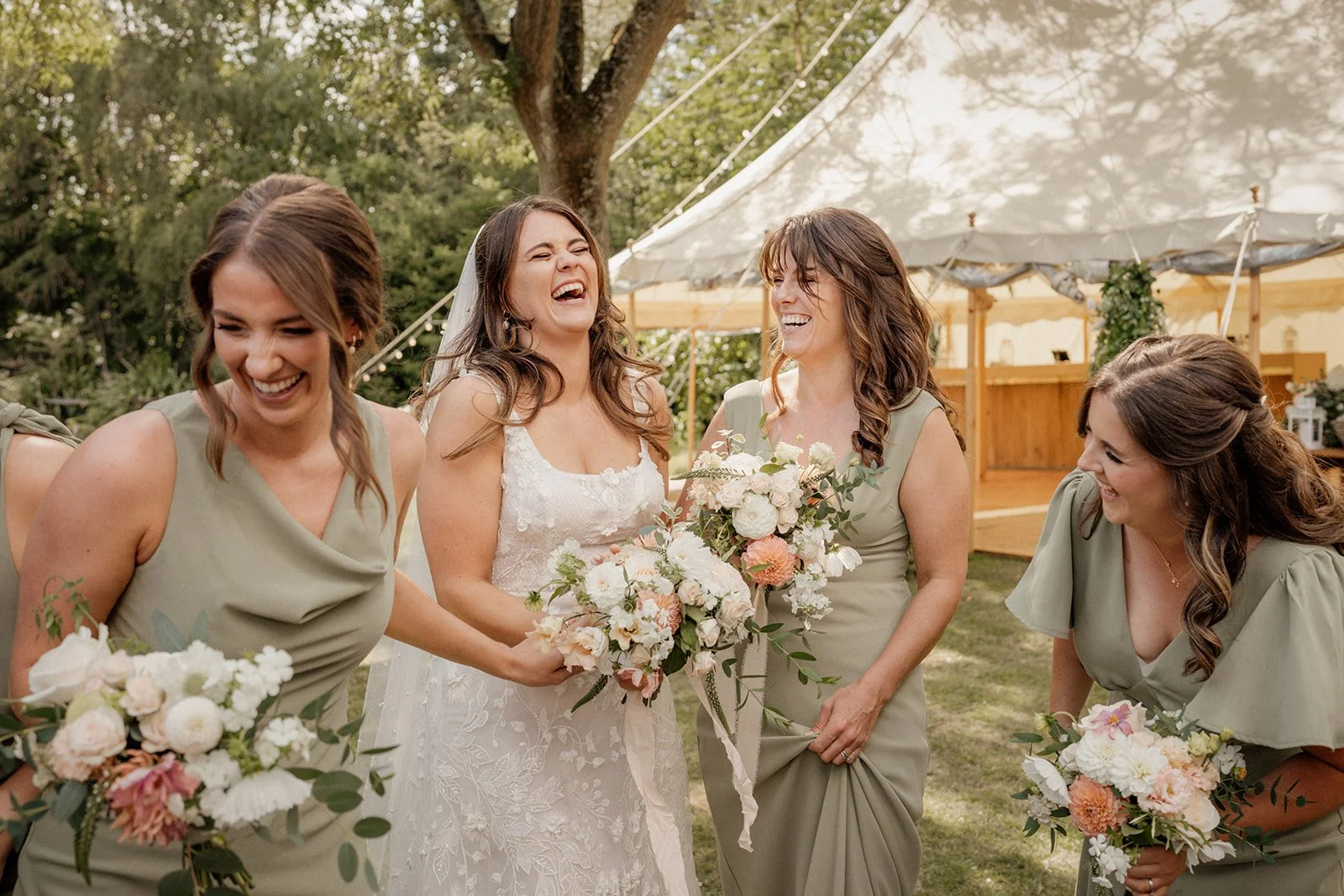 Bride and bridesmaids laughing and sharing a moment outdoors during a wedding celebration. The bride is wearing a white lace dress, and the bridesmaids are in matching sage green dresses, each holding bouquets of flowers. They are under a large white