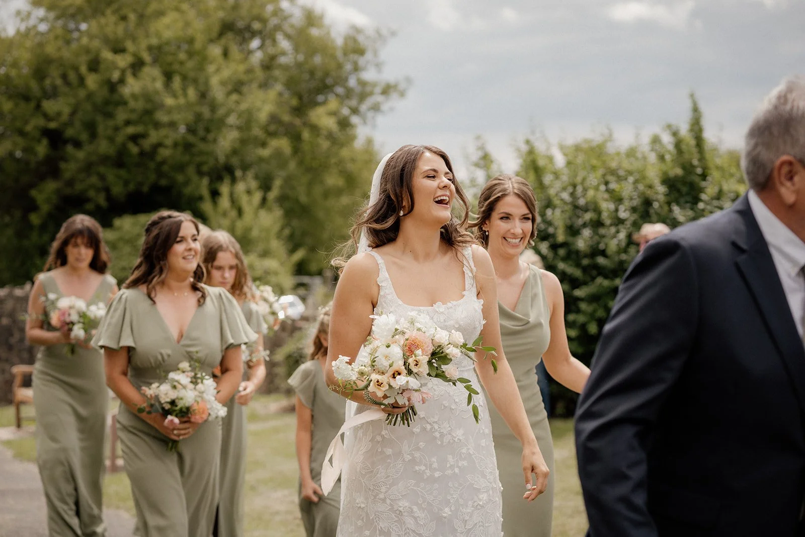 A bride in a white lace wedding dress holding a bouquet of flowers, smiling and walking outdoors surrounded by bridesmaids in matching light green dresses during a wedding ceremony.