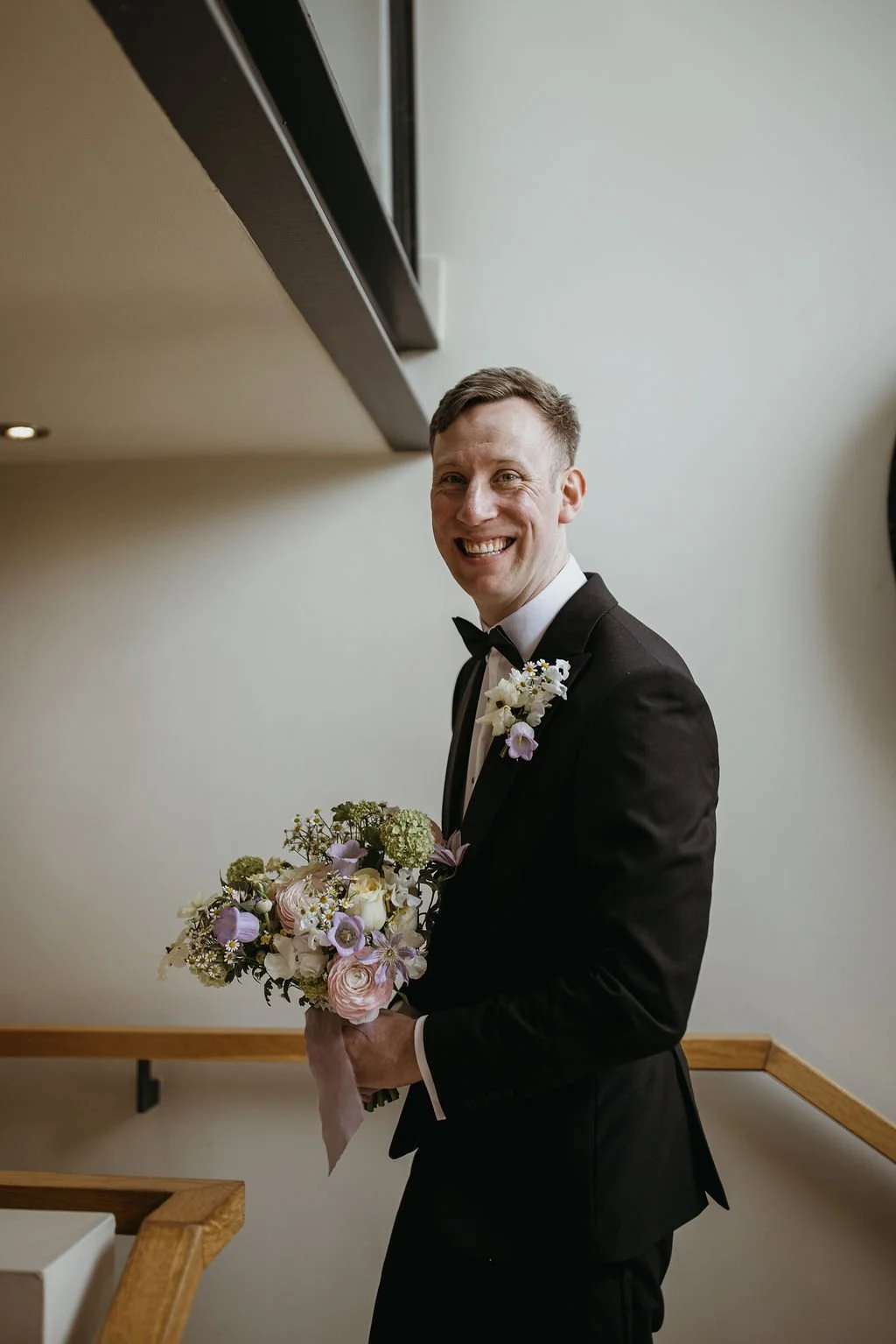 A smiling man in a black tuxedo and bow tie holding a bouquet of flowers, standing indoors against a plain light-colored wall.