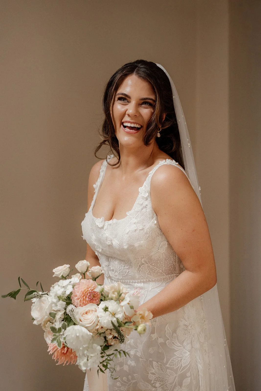 A smiling bride in a white wedding dress with lace details, holding a bouquet of pink and white flowers.
