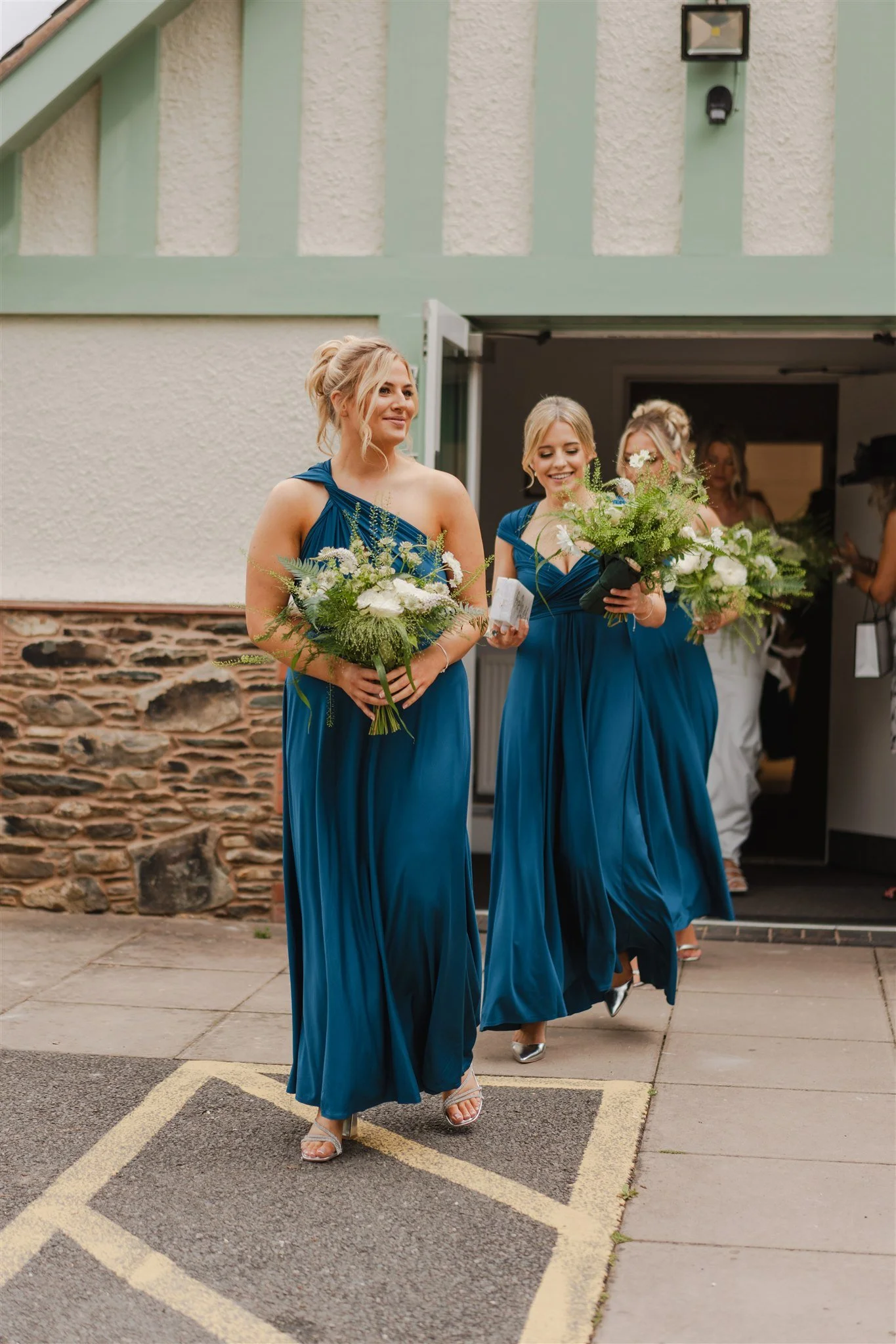 Bridesmaids in long blue dresses holding bouquets walking out of a building.