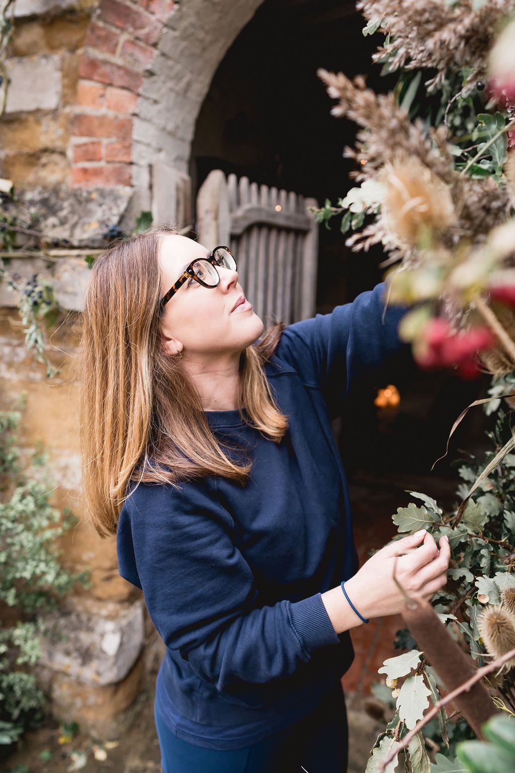A woman with brown hair and glasses tending to plants or flowers on a garden wall.