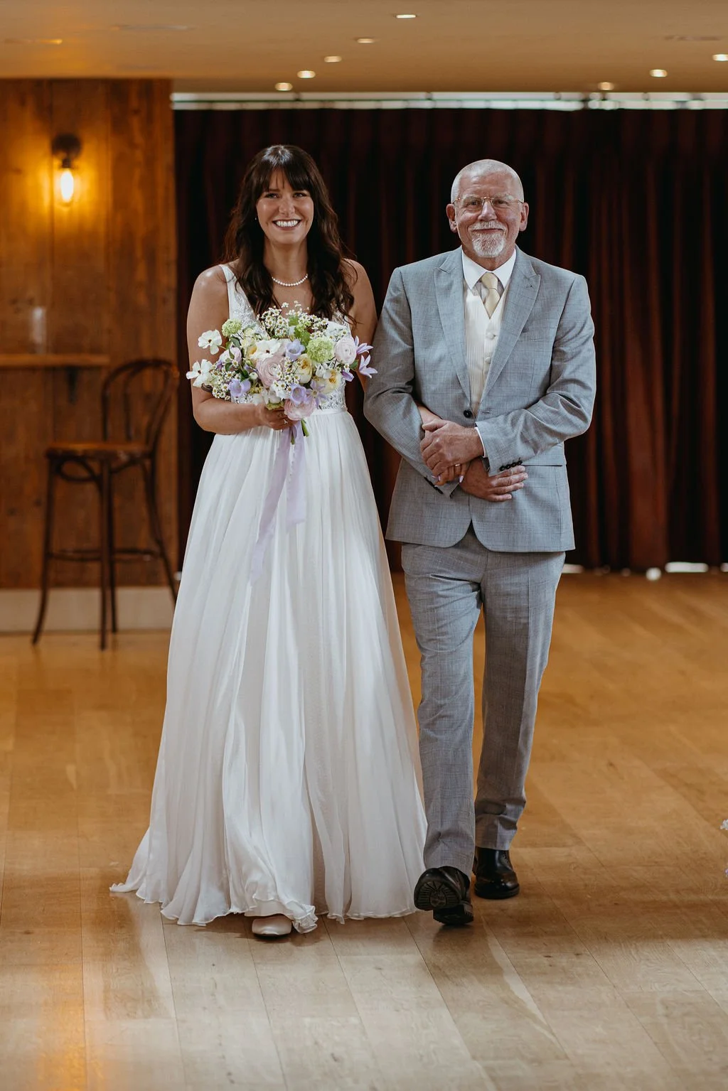 A bride in a white wedding dress holding a bouquet of flowers walking down the aisle with a man in a light gray suit at a wedding ceremony in a wooden indoor venue.