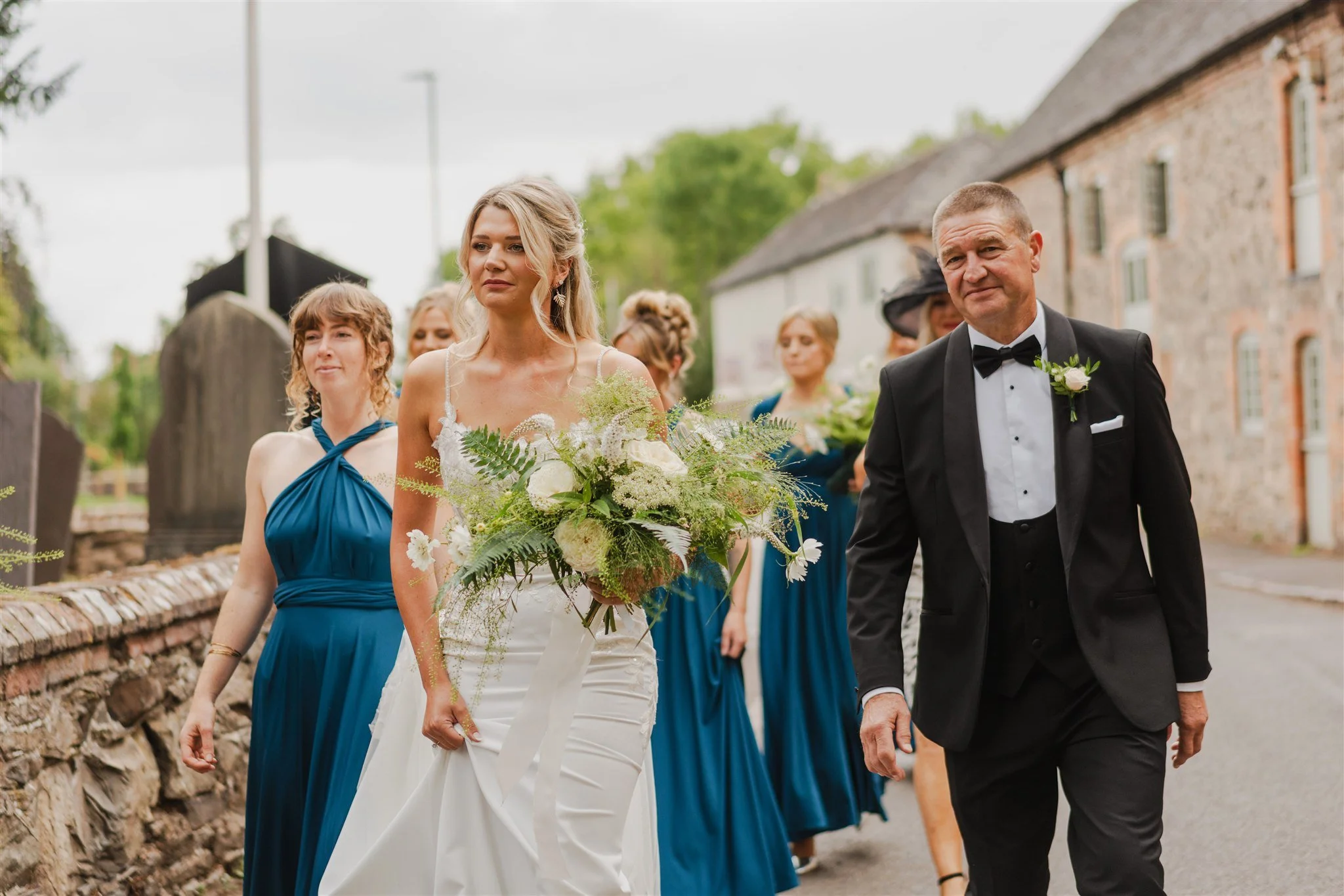 A bride in a white wedding dress walking with a man in a black tuxedo, surrounded by bridesmaids and guests outdoors.