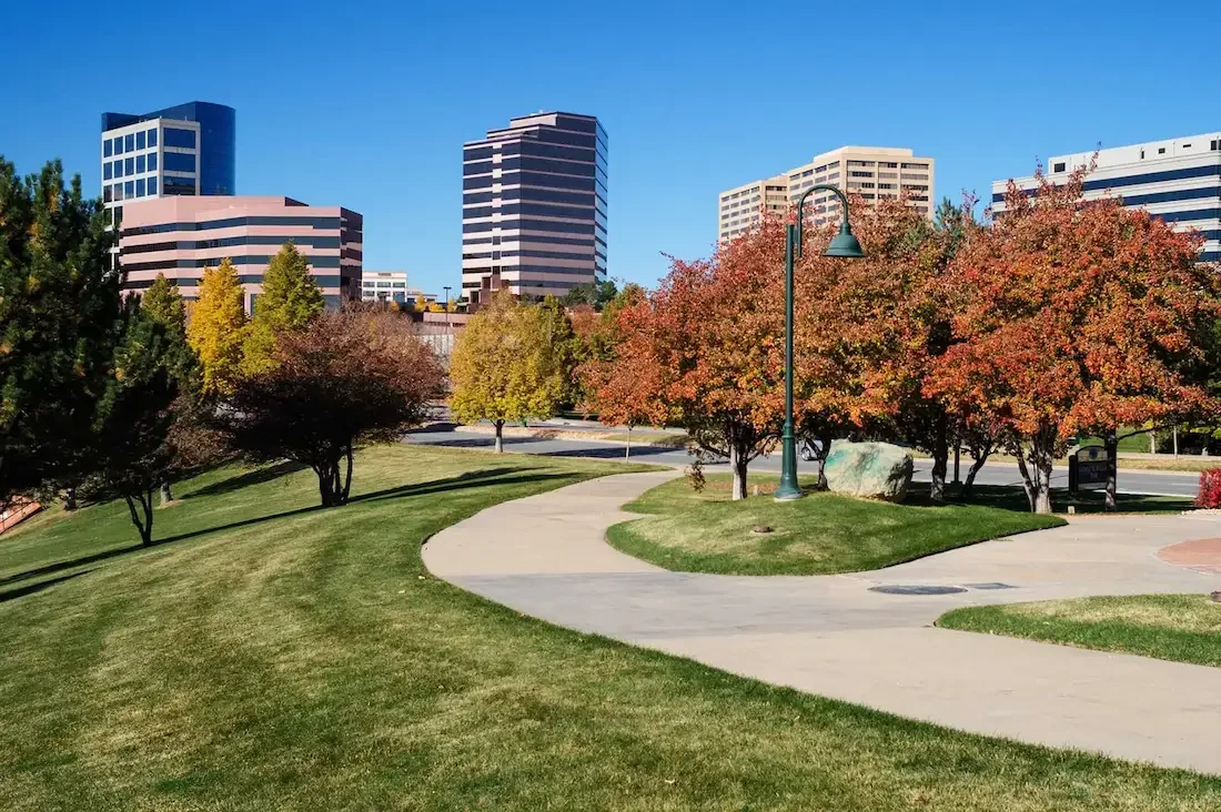 A park with green grass, trees with colorful autumn leaves, and a curved concrete pathway. In the background, there are tall modern office buildings under a clear blue sky.