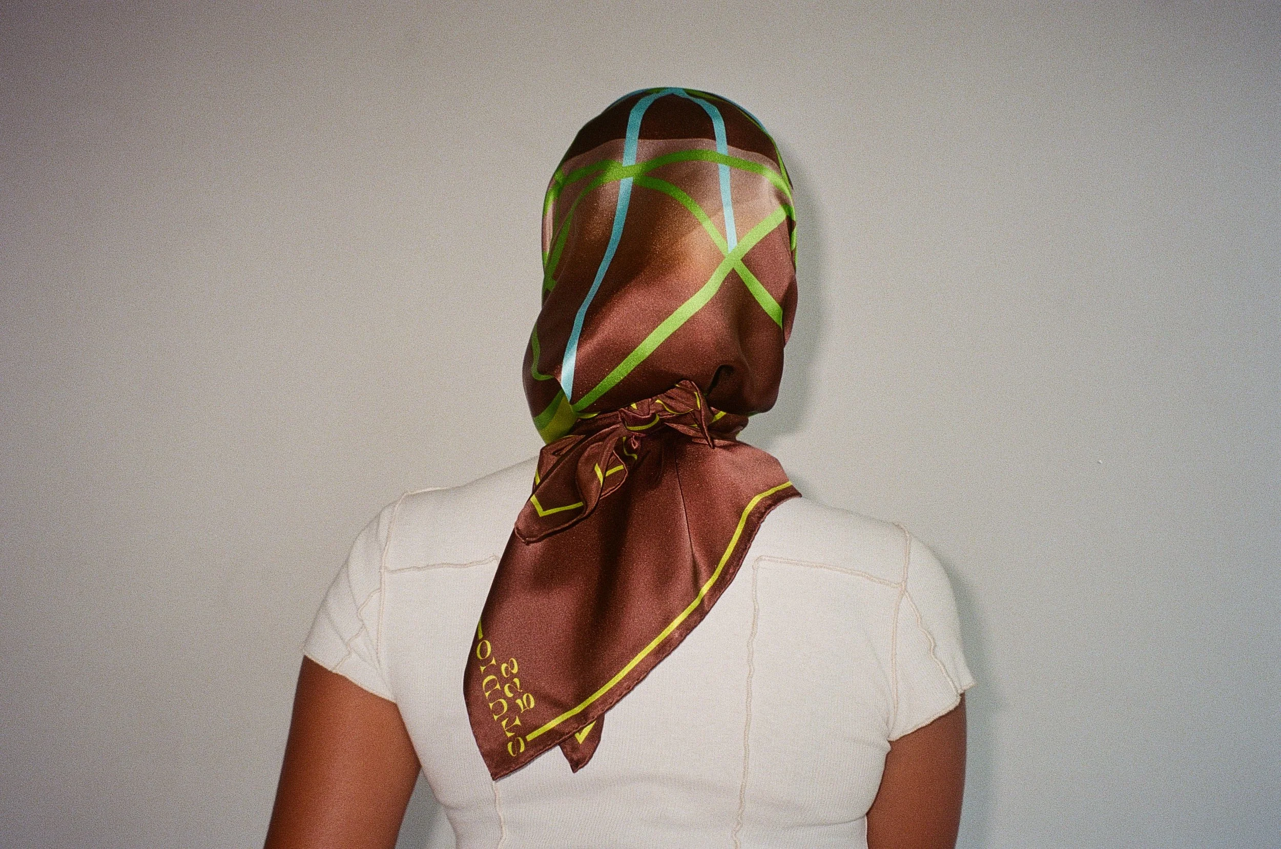 Person wearing a headscarf with green, blue, and tan patterns, tied at the back of the neck, and a white shirt with orange stitching.