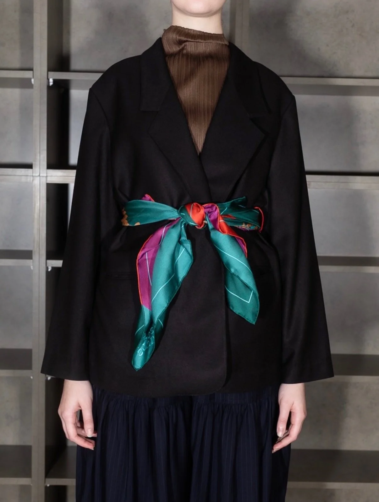 Person wearing a black blazer with a colorful silk scarf tied at the waist, brown high-neck top underneath, and dark pleated skirt, standing in front of a metal shelving unit.