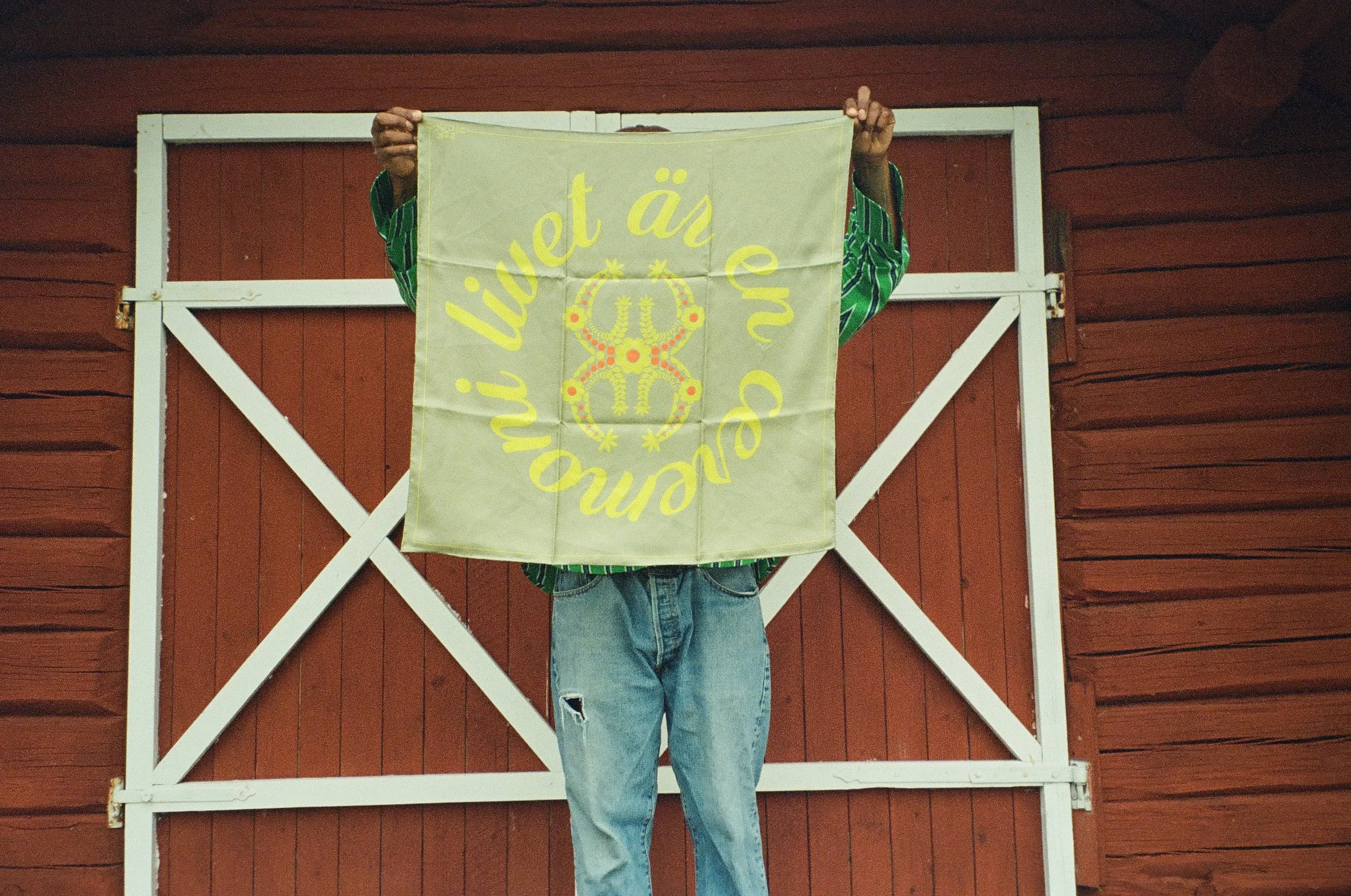 Person holding a light yellow flag with yellow text and a graphic design, standing in front of a red wooden barn with white trim, wearing jeans and a green striped shirt.