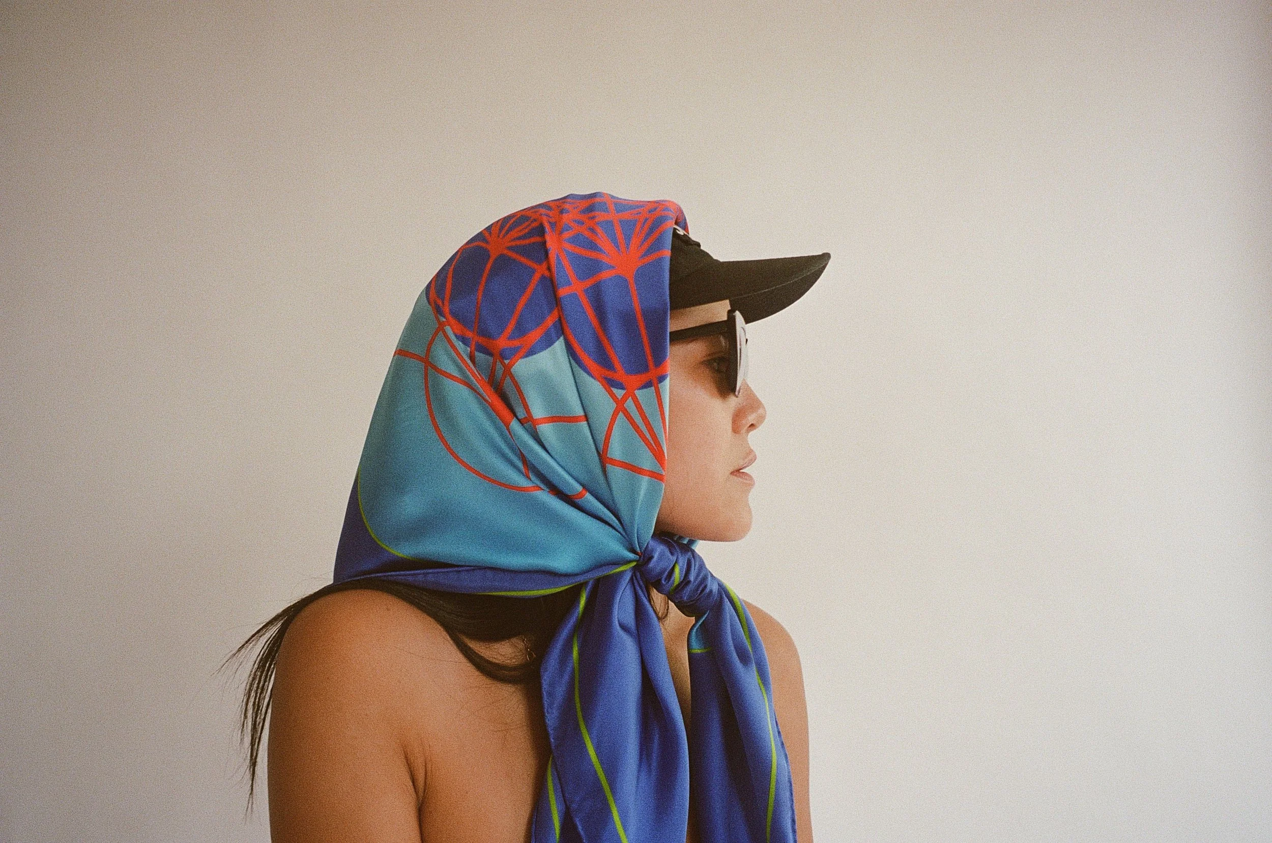 A woman in profile wearing a colorful headscarf, sunglasses, and a baseball cap, with a plain light background.
