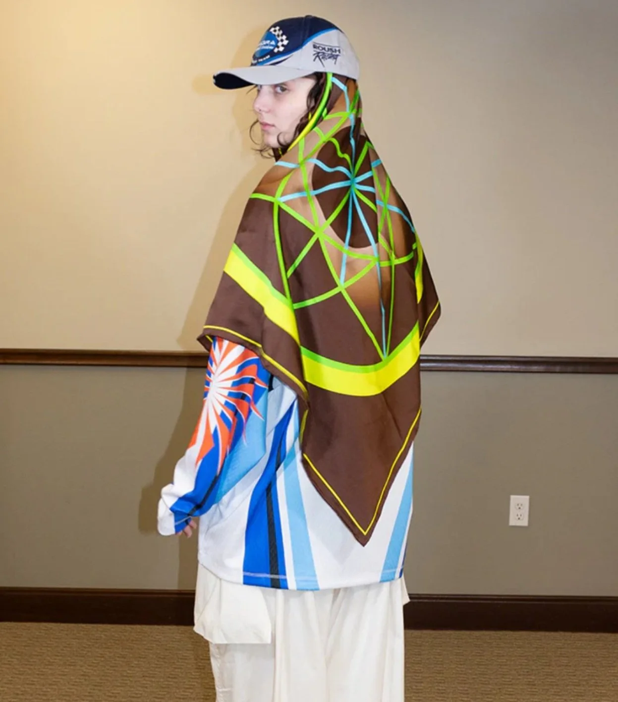 Person wearing a cap, colorful shirt, and a brown shawl with neon yellow and blue geometric patterns over their shoulders, standing in a room with beige walls and carpet.