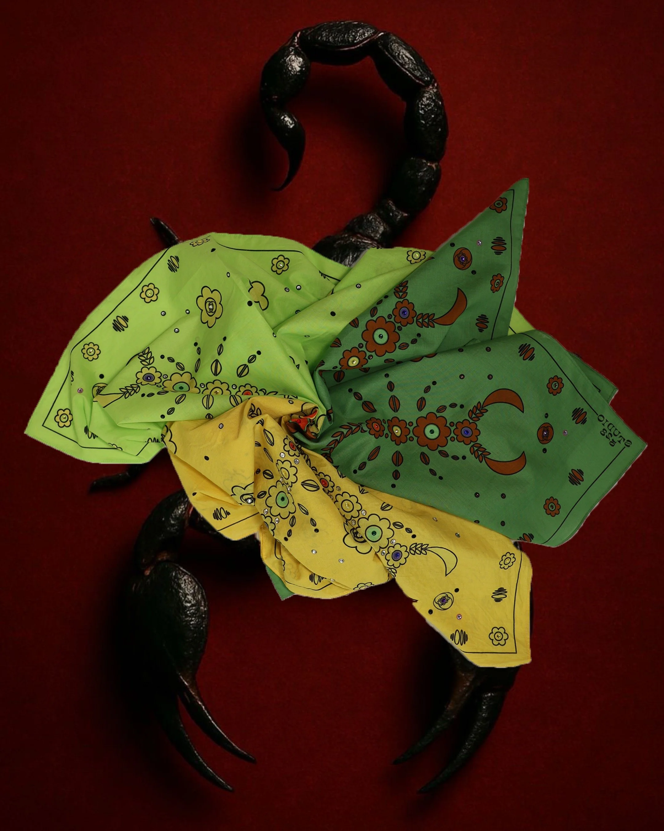 A black scorpion with a shiny exoskeleton wearing a green and yellow bandana with floral designs on a red background.
