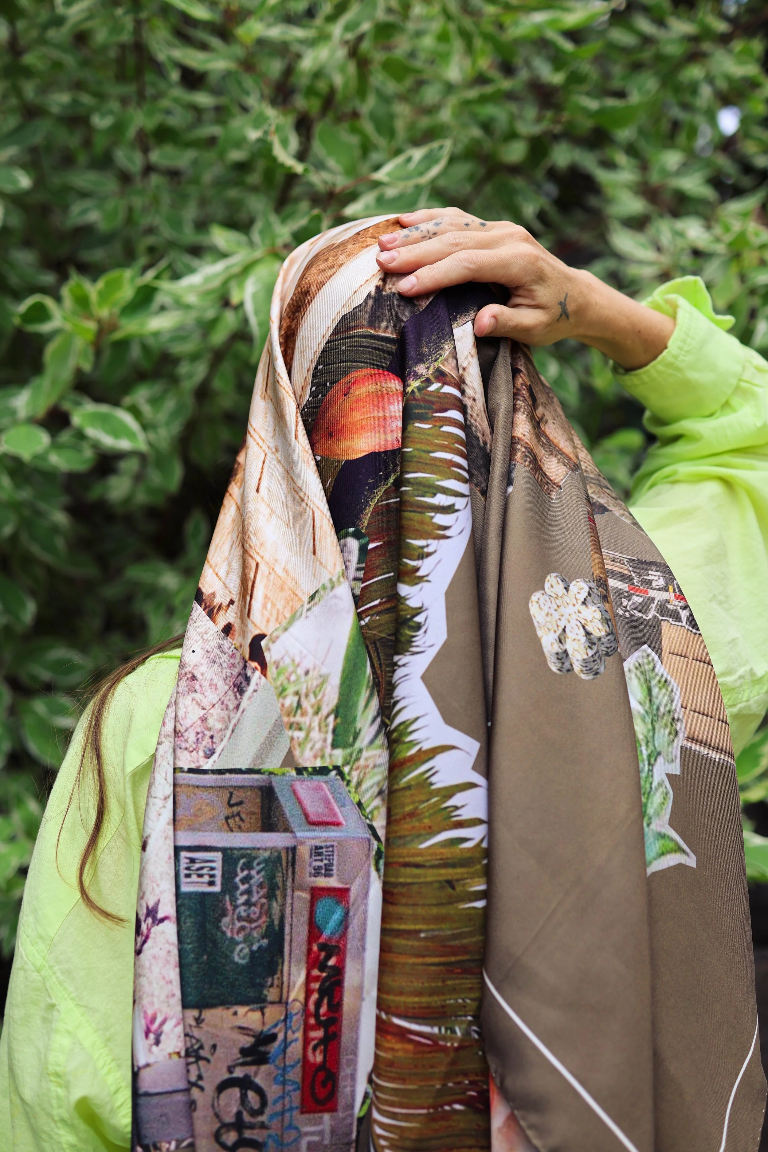 Close-up of a person's hand resting on their knee, wearing a bright yellow jacket, with a tattoo on the wrist, in front of dense green foliage. The person has tattoos on fingers and wrist, and a scarf with a map and magazine images.