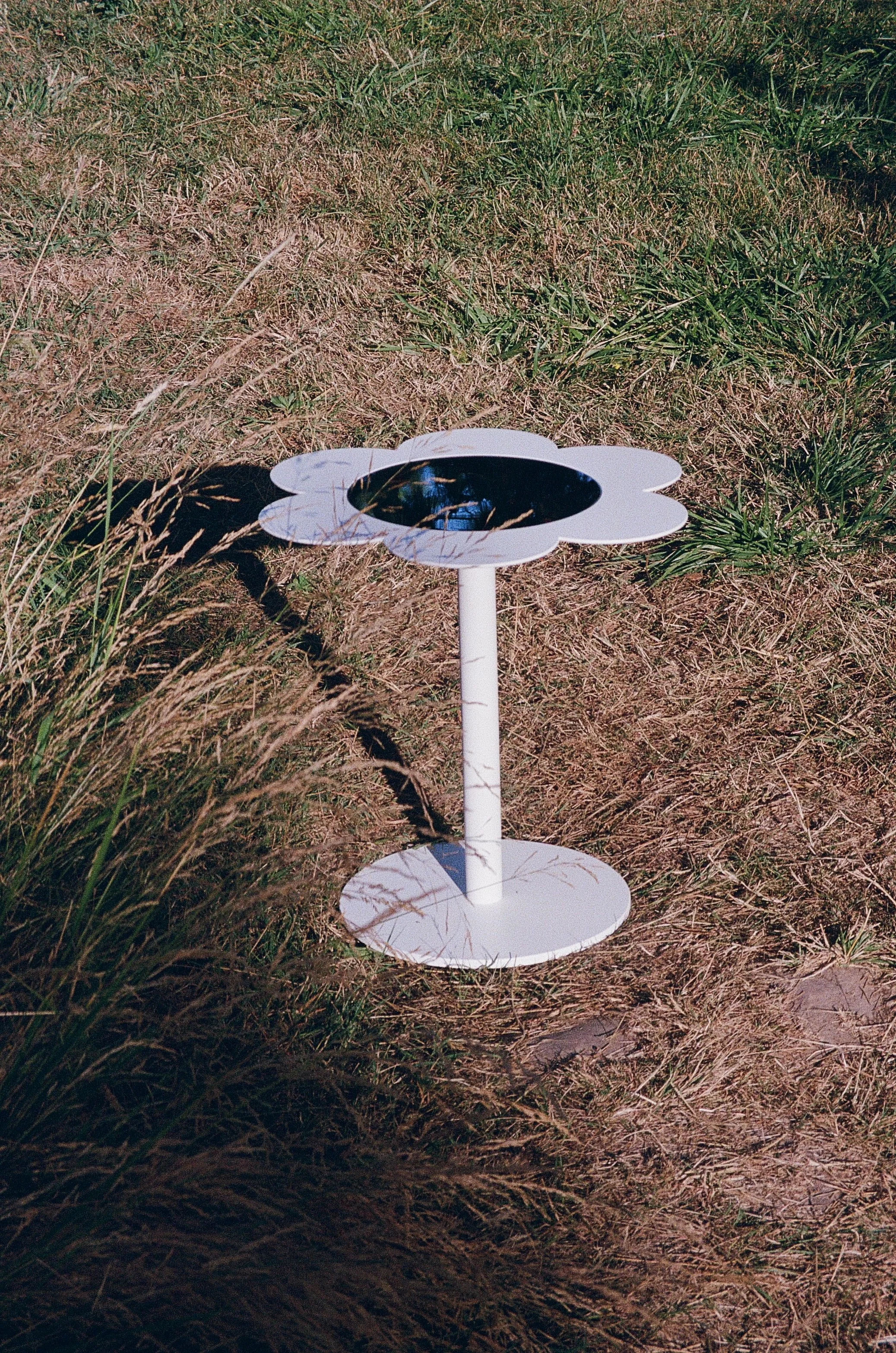 A white flower-shaped side table with a mirrored glass top,  situated outdoors on a patch of grass and dirt.