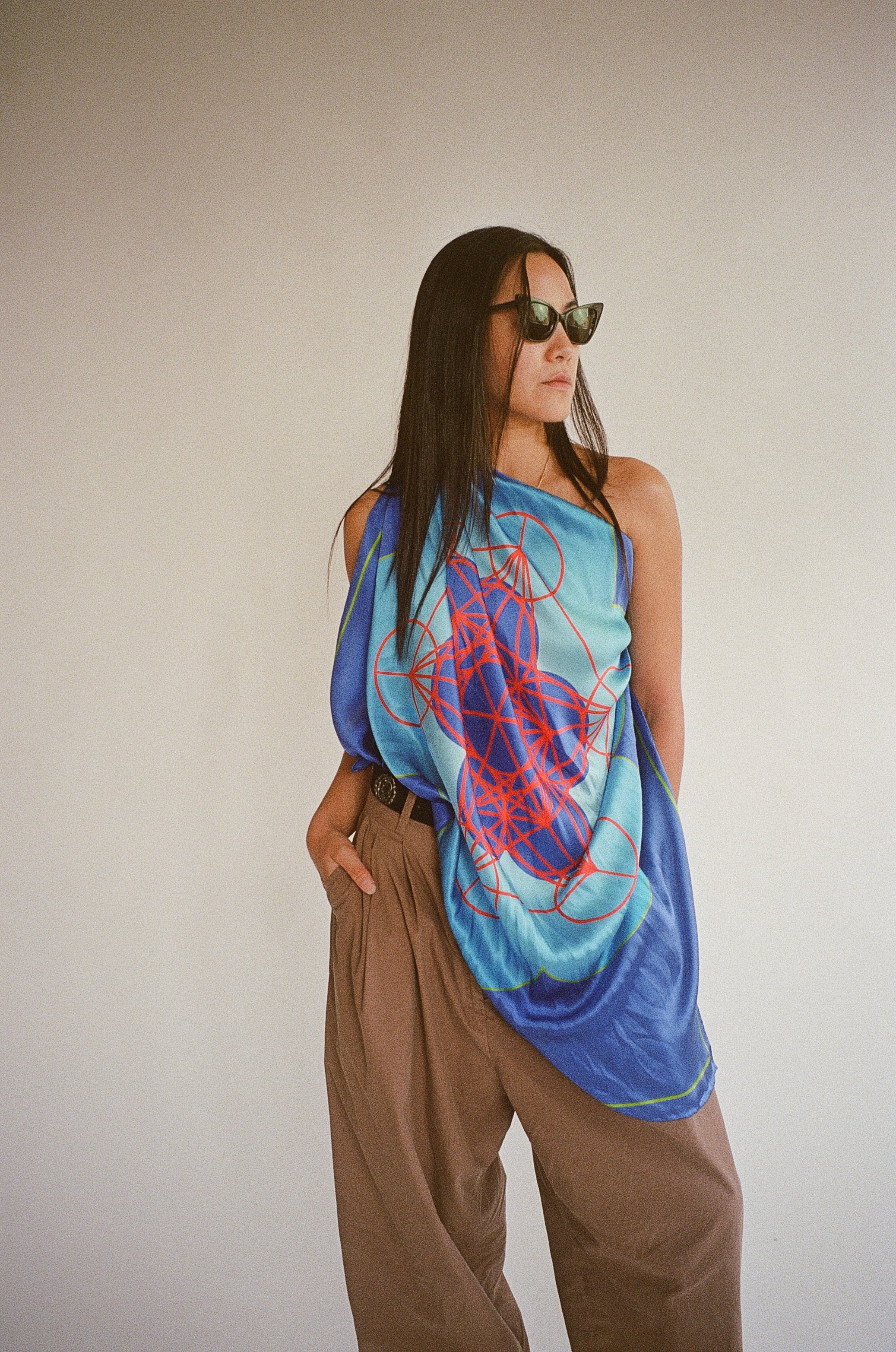 A woman with long dark hair wearing sunglasses, a colorful patterned sleeveless top, and khaki pants stands against a plain background.