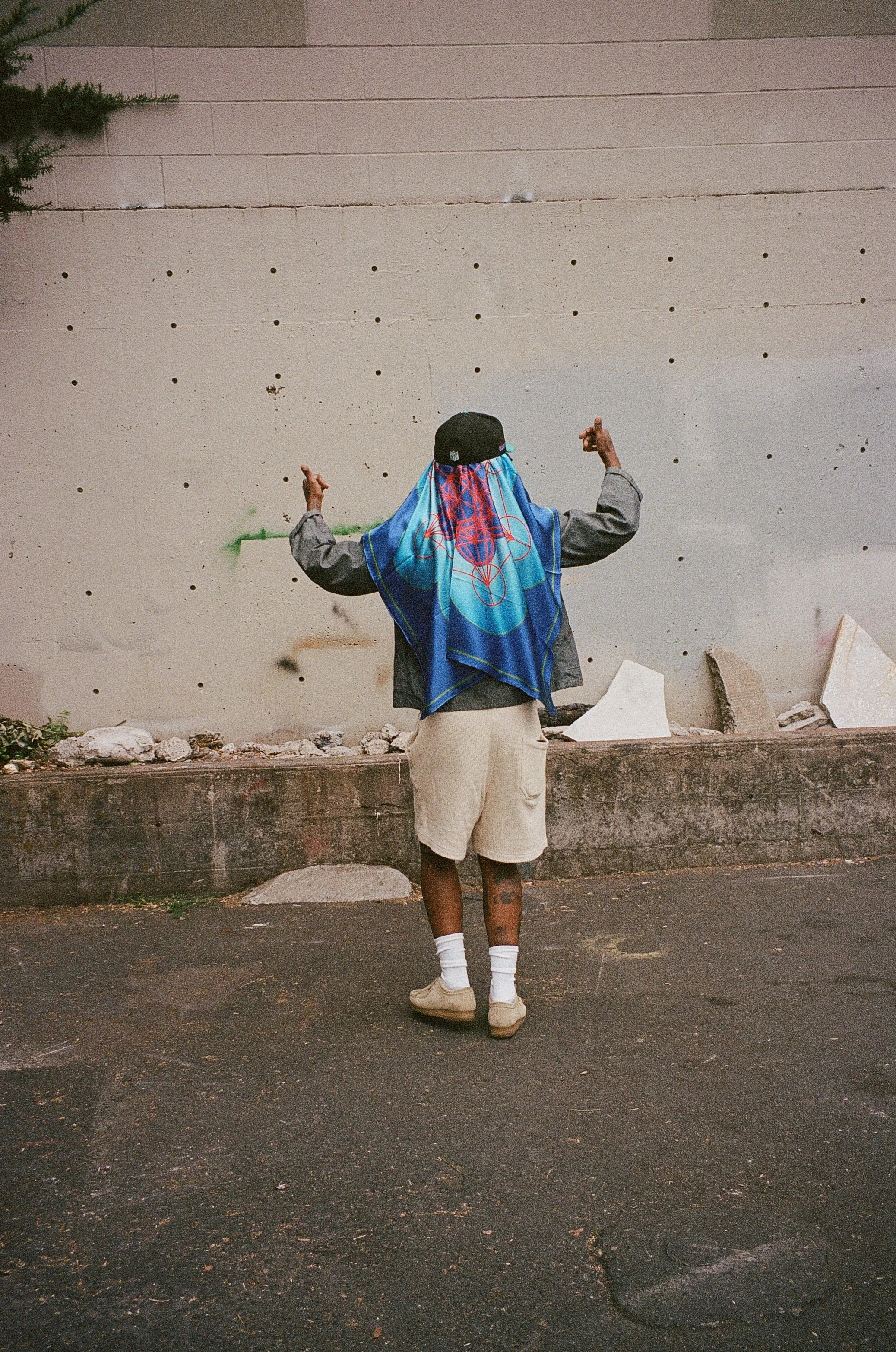 A person standing outdoors with their back to the camera, wearing a large blue and red patterned headscarf, a black cap, gray jacket, beige shorts, white socks, and beige shoes, gesturing with both middle fingers raised, in front of a light-colored wall with multiple small holes and some debris on the ground.