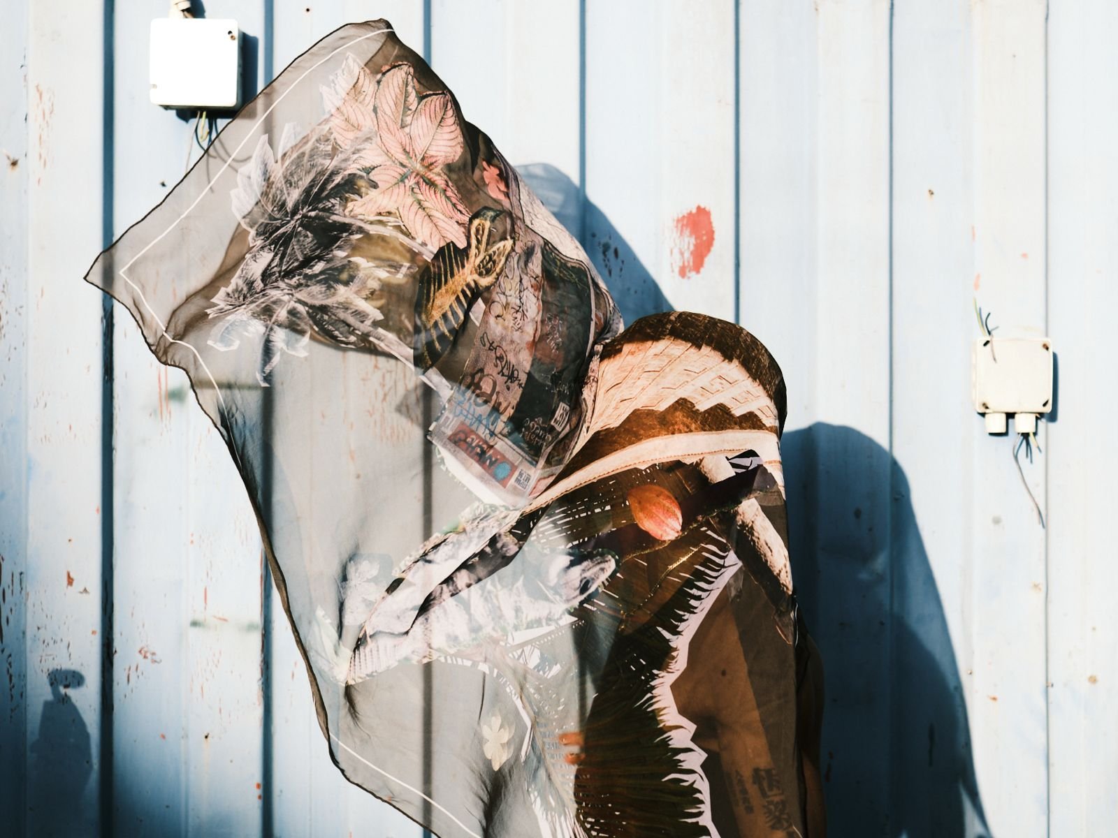 A person wearing a transparent jacket with floral and animal print patterns leans against a weathered light blue wall with an electrical box and wiring.