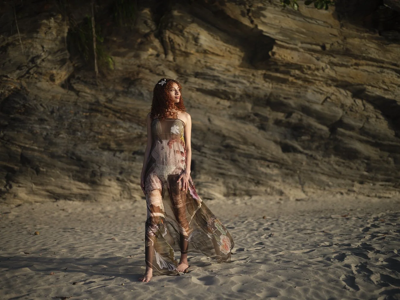 A woman with curly red hair standing barefoot on sandy beach at sunset, wearing a sheer silk scarf that is used as a dress, with a rocky cliff in the background.