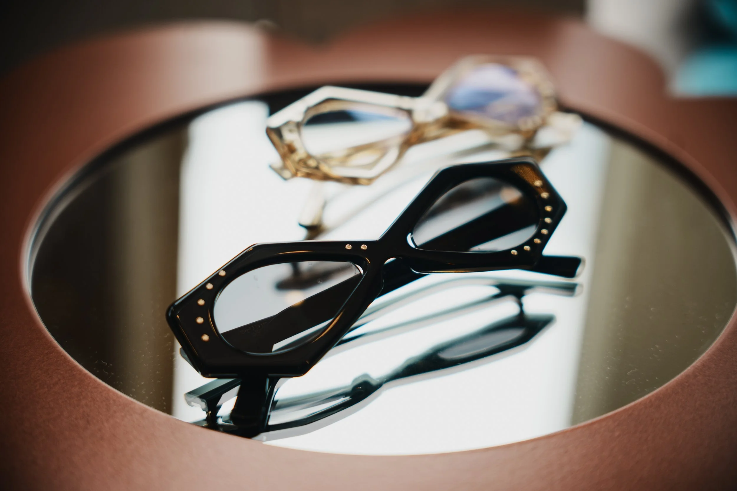 Close-up of a pair of black glasses reflected in a mirror with jewelry, including a gold ring, on a white surface.