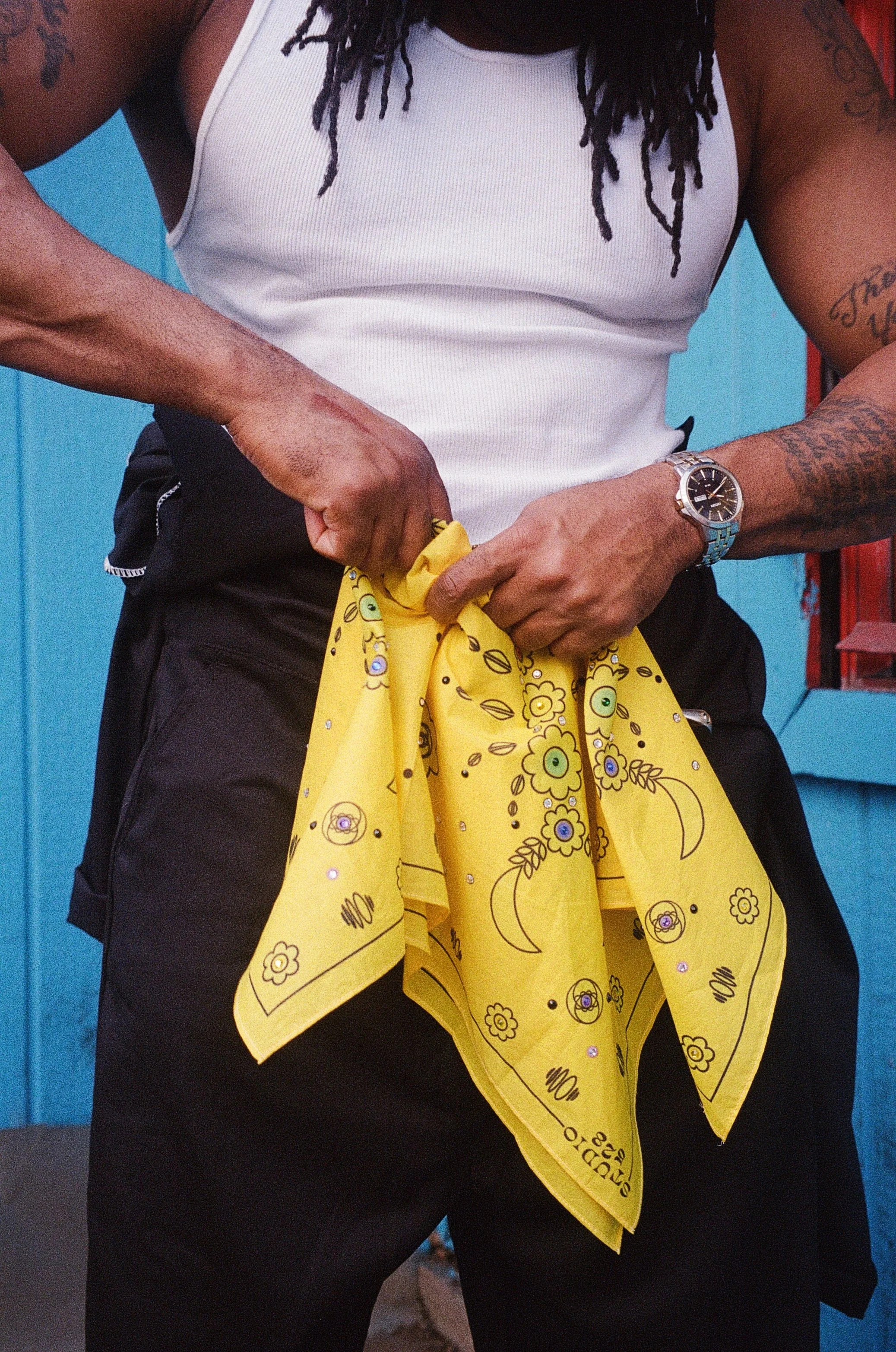 Person wearing a white tank top, black pants, and a yellow bandana with floral and moon patterns, standing in front of a blue background, with tattoos on their arms and a watch on their left wrist.