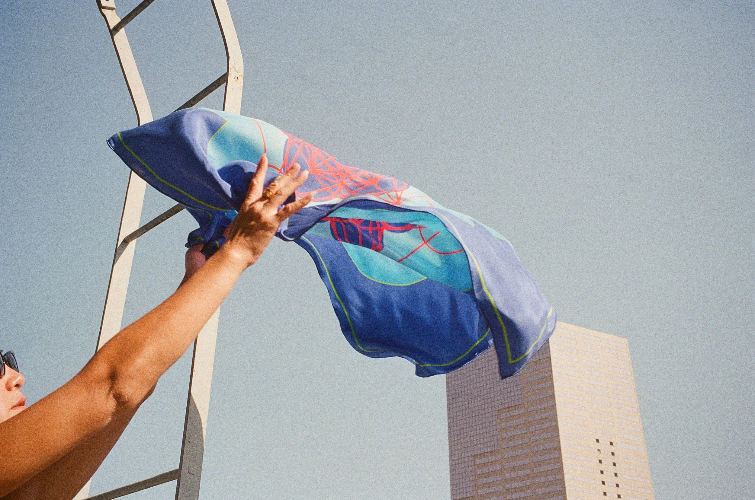 A person with a tan complexion is holding a brightly colored piece of fabric or clothing, possibly a shirt, up against a clear sky. The fabric features a vibrant geometric design with shades of blue, red, and light blue. In the background, there's a modern high-rise building with a light-colored, patterned facade and a blue sky.