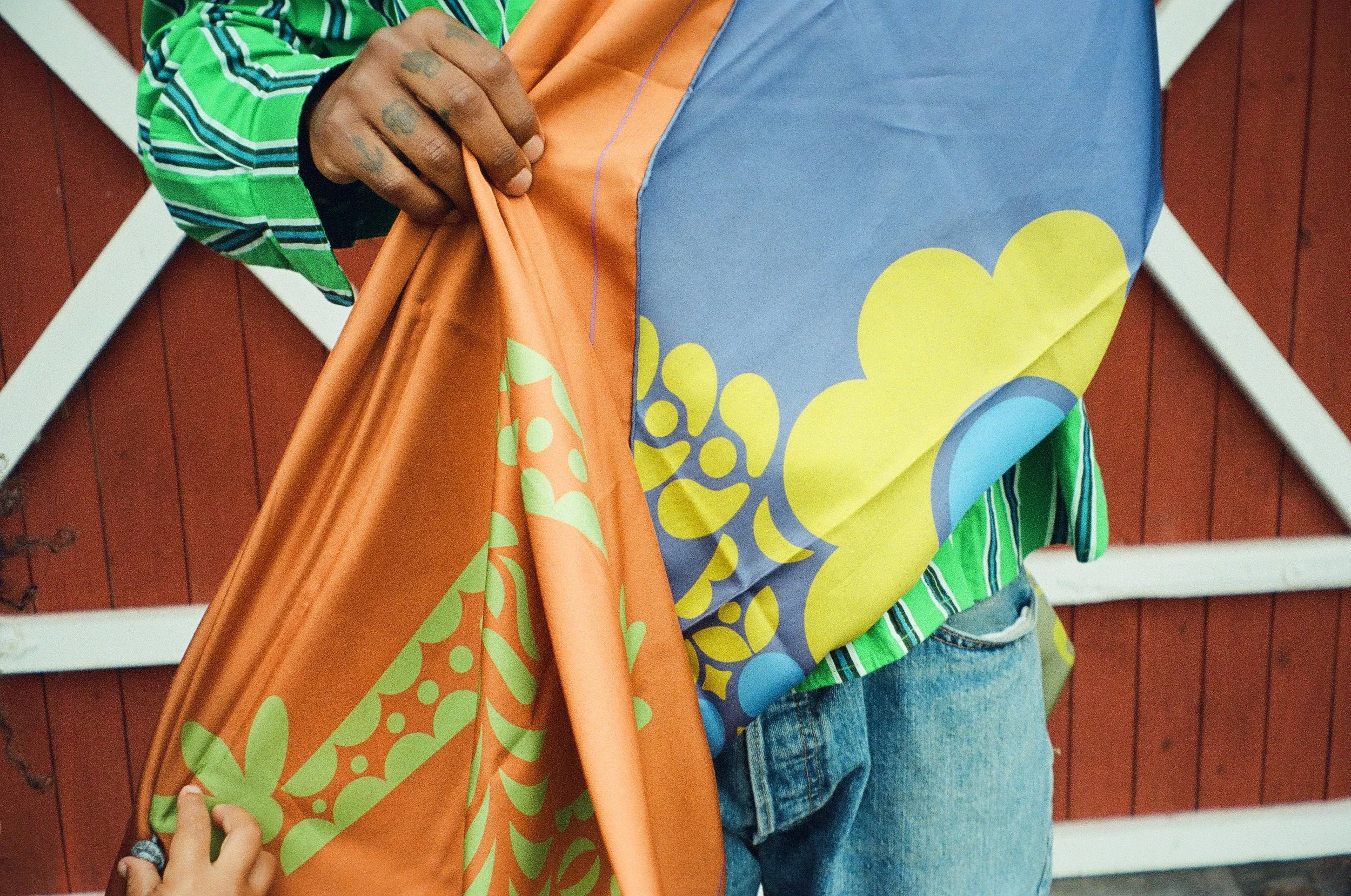 A person wearing a colorful, patterned shirt and blue jeans holding a large, vibrant orange and blue printed fabric or paper in front of a red wooden wall.