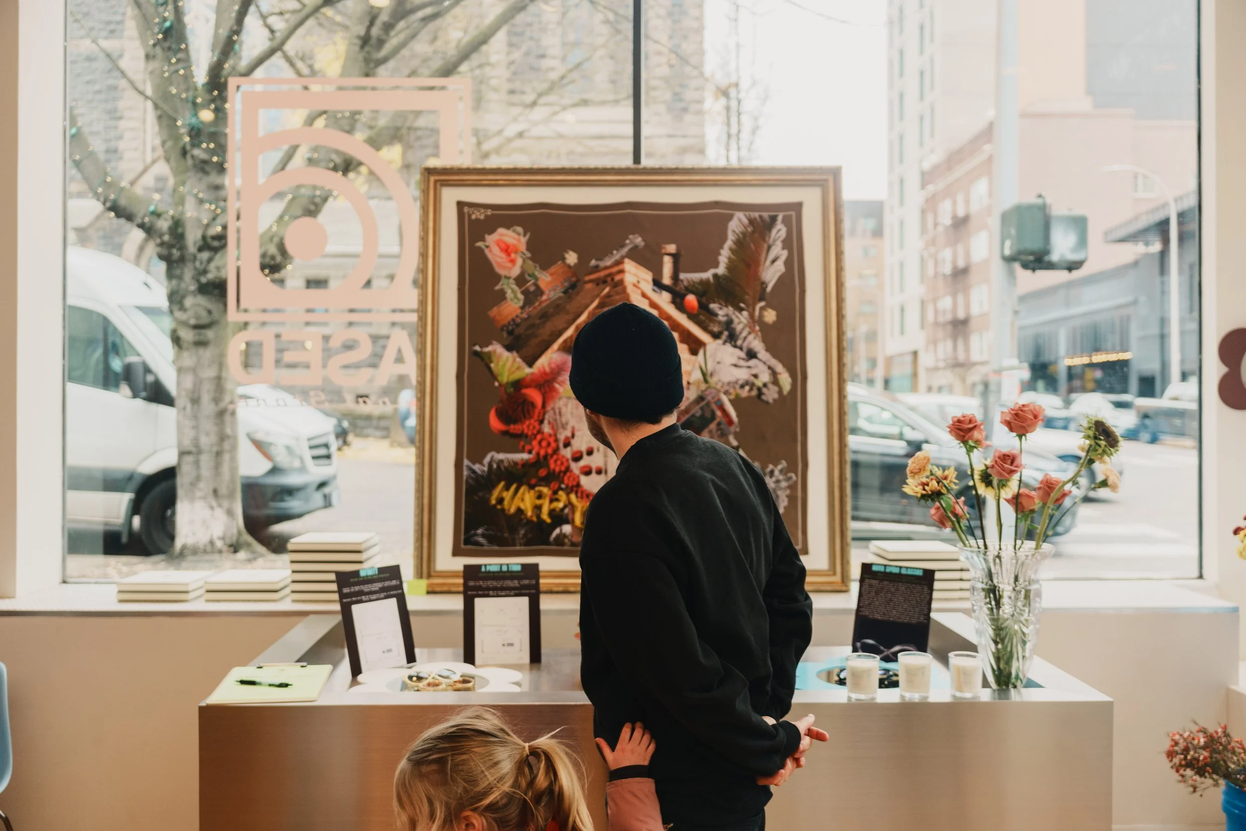 Inside an art gallery with a man and a young girl. The man is looking at a large framed artwork depicting a floral arrangement on a table. The girl is reaching out towards the artwork. Outside the window, there are trees and city buildings. The gallery window has a Facebook logo on the glass.