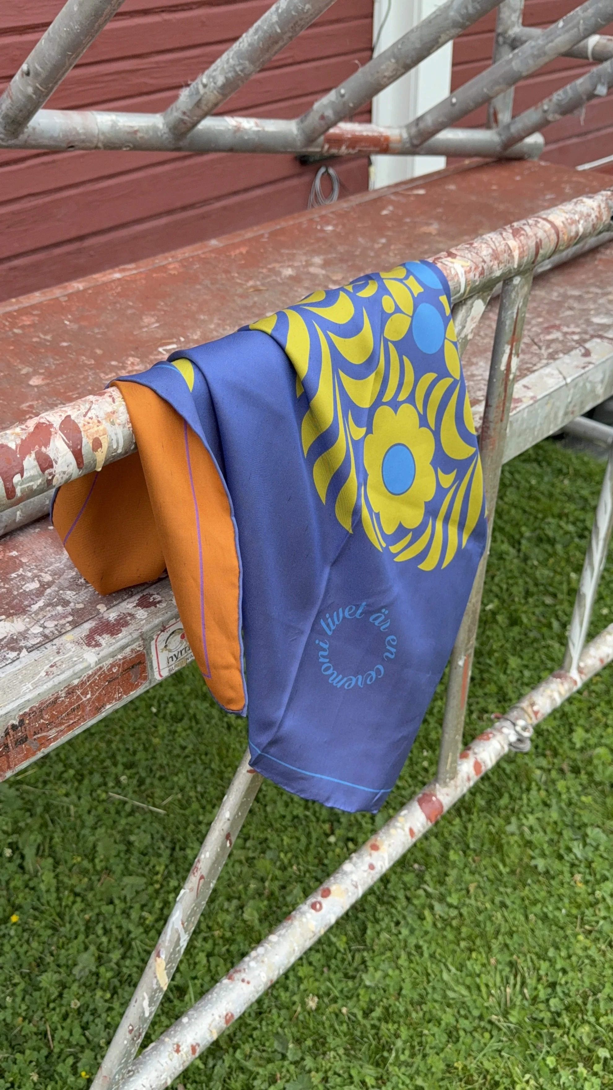 A colorful cloth with blue, orange, yellow, and green patterns hanging over a paint-splattered metal scaffold outside, with grass visible below.