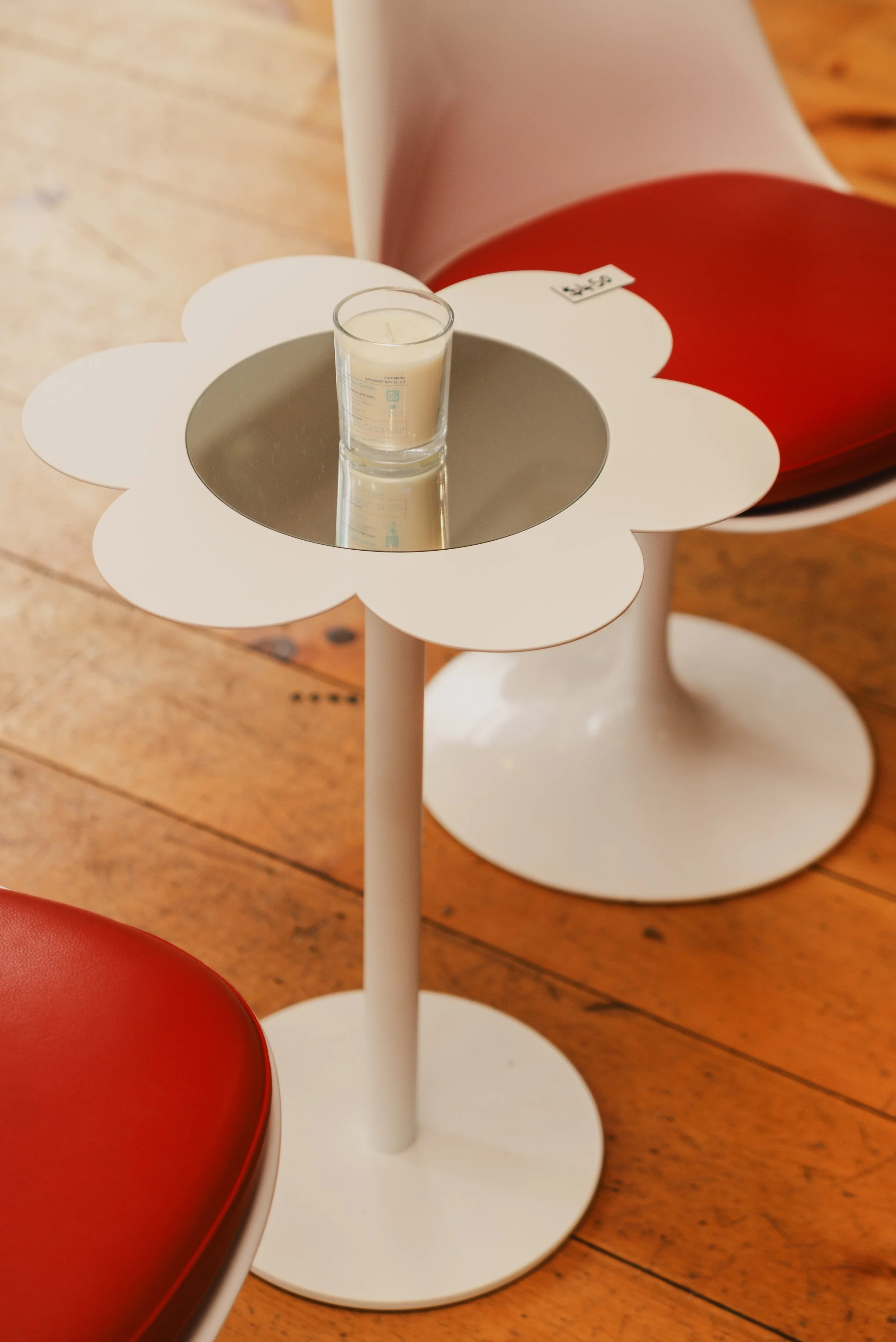 A white flower-shaped side table with a round mirror in the center, holding a white candle in a glass container. Part of a red chair or cushion is visible in the foreground, and wooden floorboards are in the background.