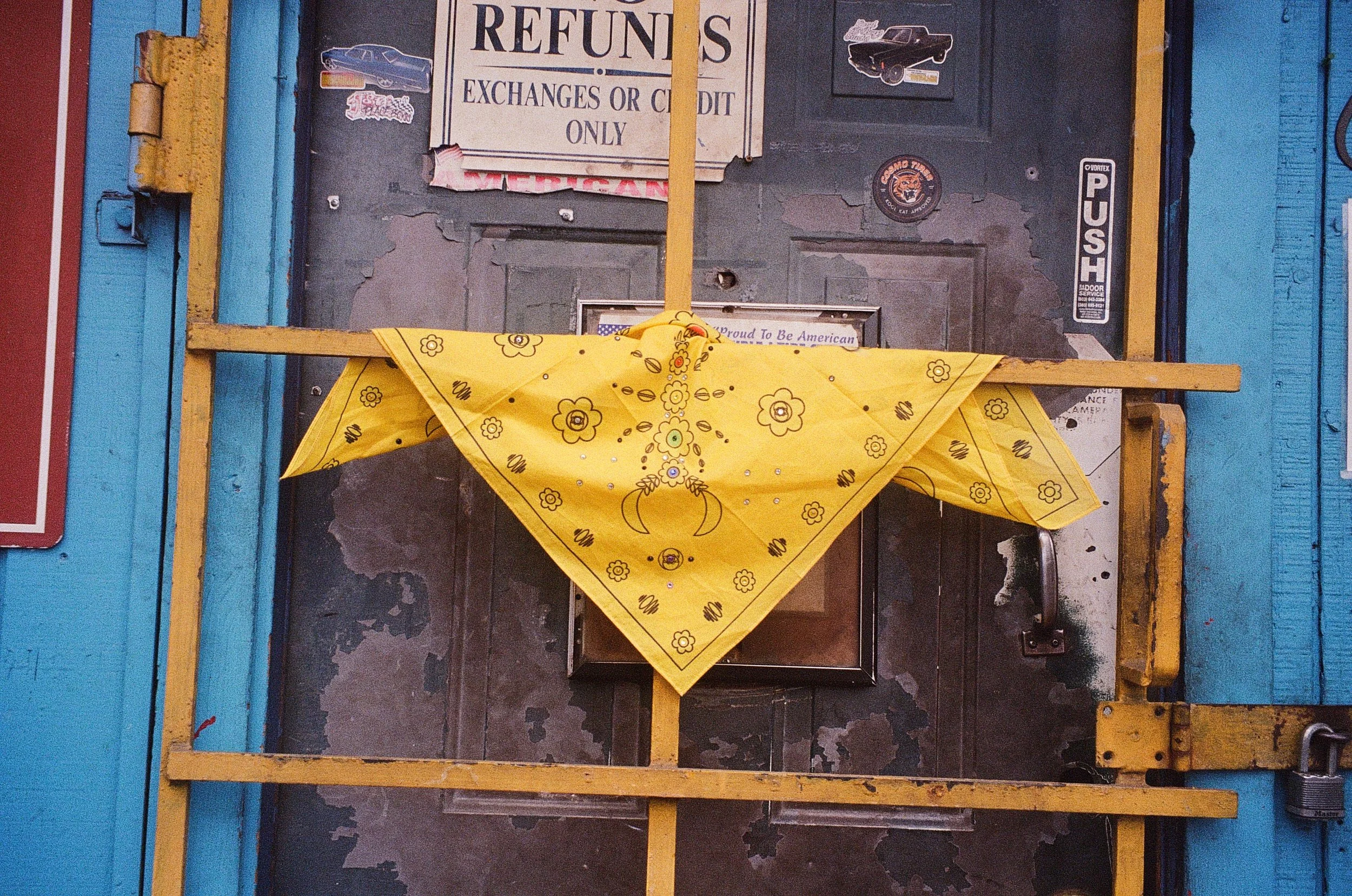 A black door with various stickers and signs, including a large 'REFUNDS' sign, a 'PUSH' sticker, and several car images. A yellow bandana with a floral pattern, folded in half diagonally, is attached to a yellow wooden frame across the door, forming a makeshift barrier or decoration.