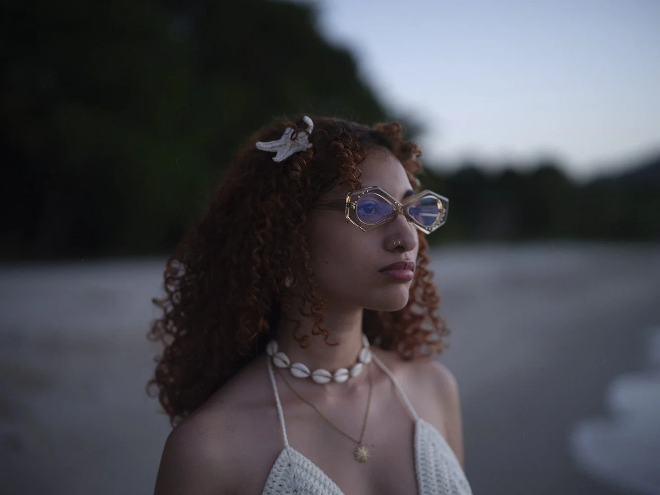 A young woman with curly red hair wearing sunglasses, a shell necklace, and a white crochet top stands on a beach during dusk, looking into the distance.