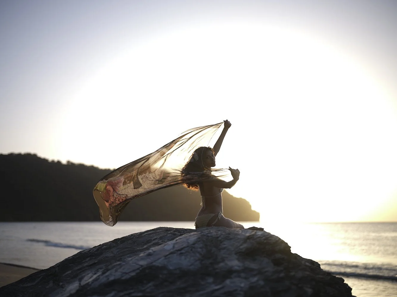 A woman sitting on a rock at the beach during sunset, holding a sheer fabric above her head.