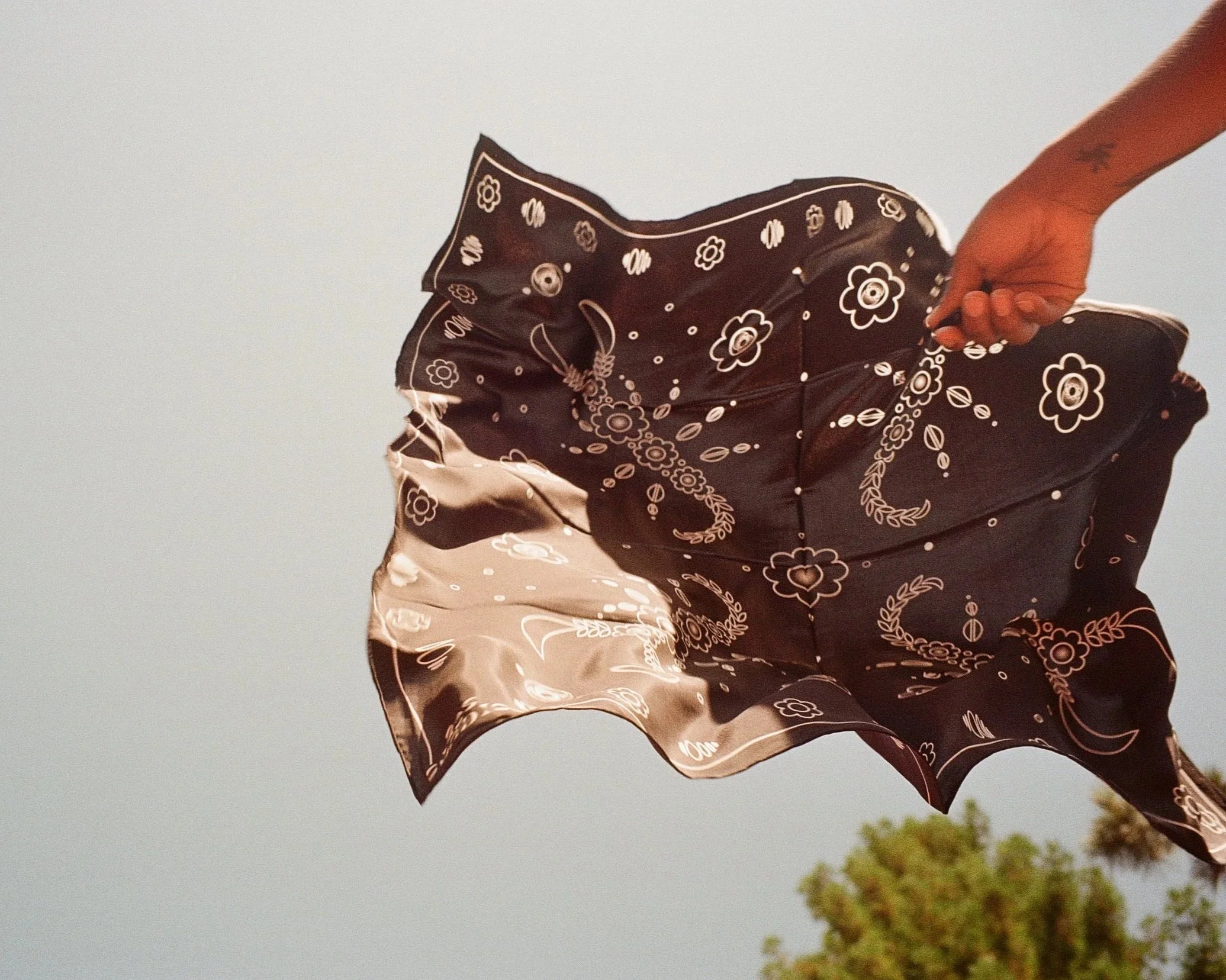 A person holding a black bandana with a white floral pattern, waving it in the air against a clear sky background.