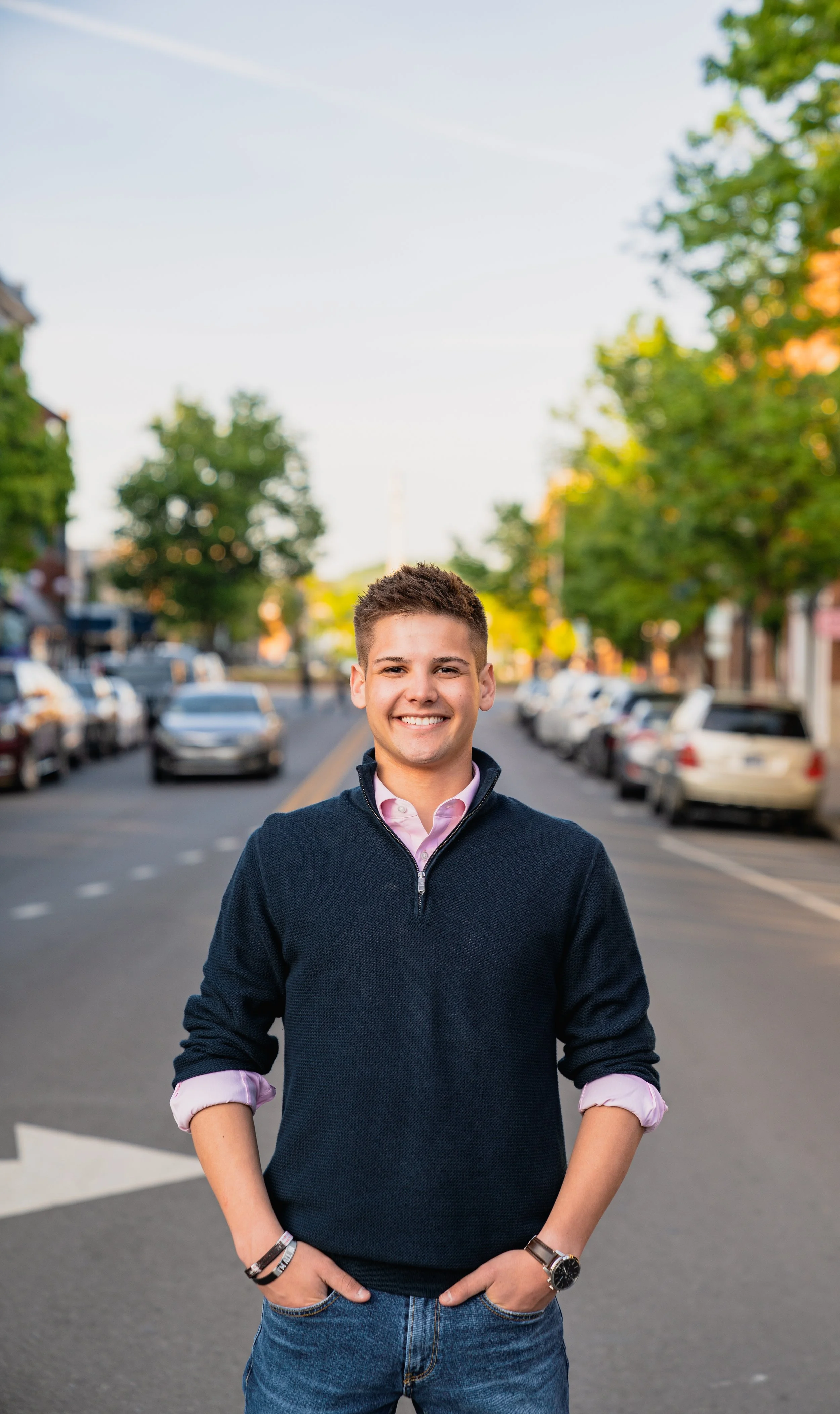 Young man wearing a navy sweater over a pink collared shirt and blue jeans, smiling with hands in pockets, standing on a city street lined with parked cars and green trees, during daytime.