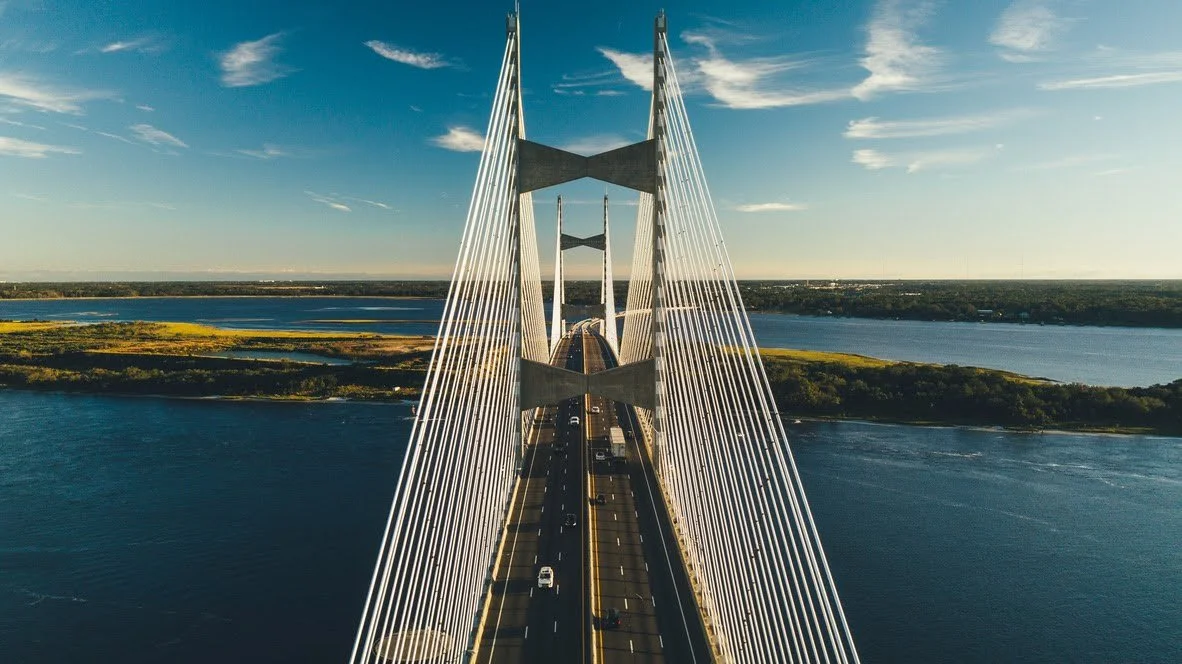 Aerial view of a cable-stayed bridge crossing a wide river with green land on both sides and a blue sky above.