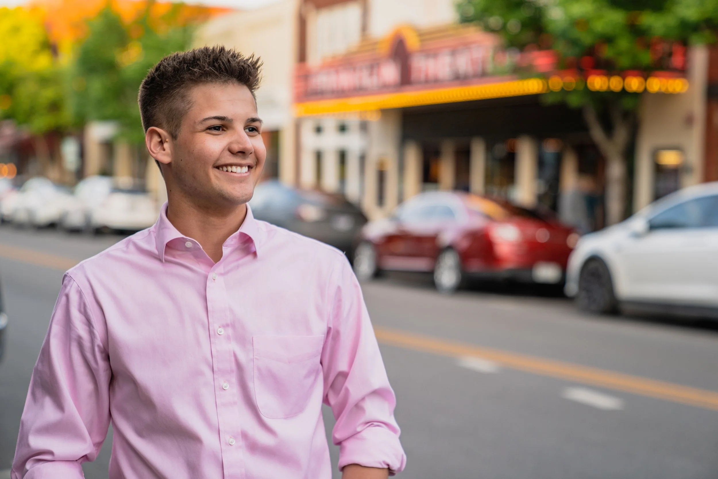 A young man smiling, wearing a pink button-up shirt, standing outdoors on a city street with cars and buildings in the background.