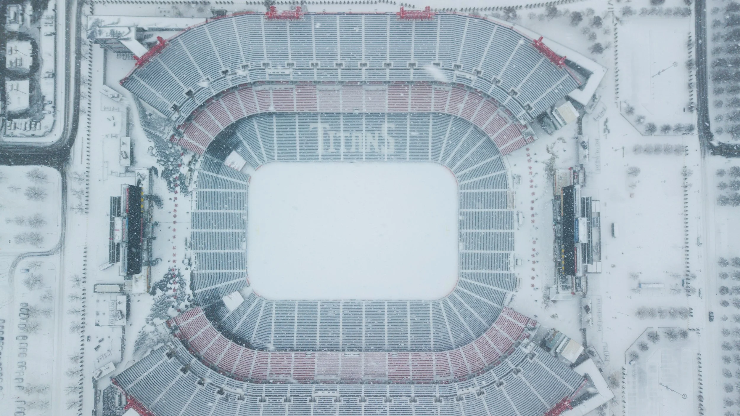 An aerial view of a snow-covered football stadium during winter, with seats and the playing field blanketed in snow.